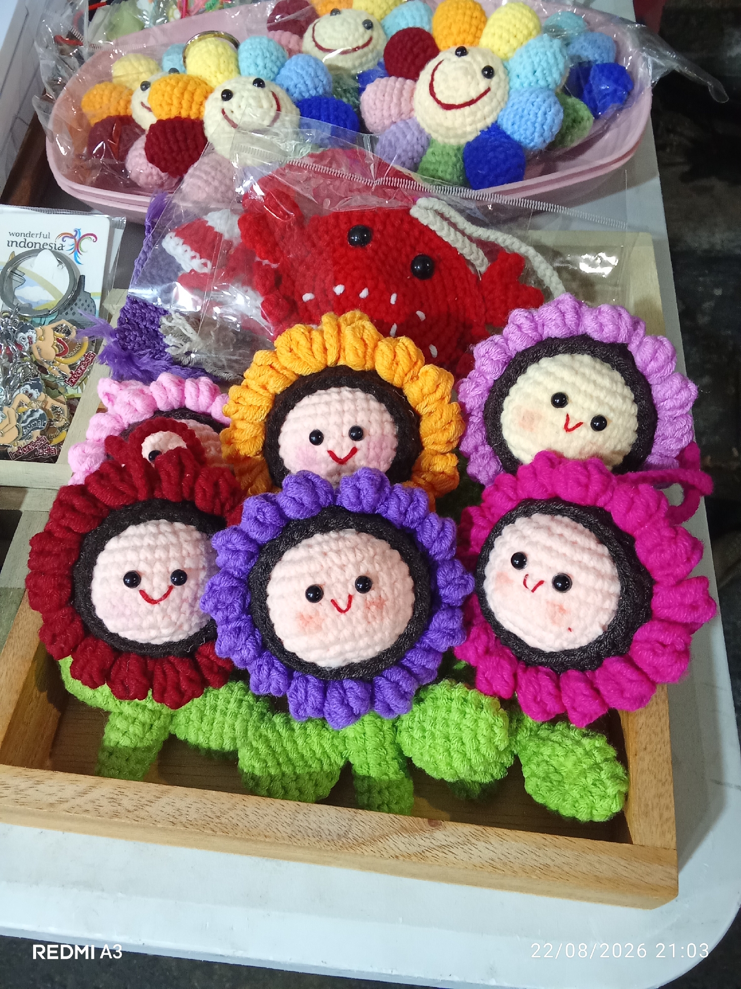 In Surakarta, Indonesia, a close-up, overhead shot reveals a wooden tray filled with crocheted items. Dominating the foreground are several cheerful, crocheted sunflowers with smiling faces, their petals in shades of red, pink, purple, and orange. Behind these, a red crocheted strawberry-like object is visible, and further back, a pink bowl overflows with colorful crocheted balls, some with smiley faces. To the left, a small stack of postcards or tags, one featuring the words "wonderful Indonesia," is partially visible, along with a keychain. The overall scene is brightly lit, suggesting an indoor setting, possibly a market stall or a craft display.