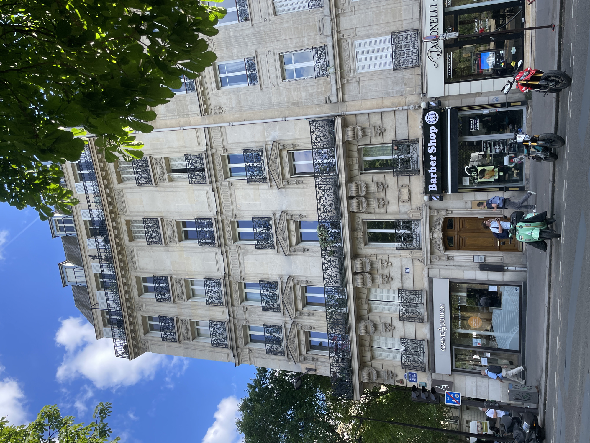 This image captures a vibrant street scene in Paris, France, on a bright, sunny day with a blue sky and scattered white clouds. The central focus is a multi-story building of classic Parisian architecture, constructed from light-colored stone. Its facade features numerous windows, many with white shutters and ornate black wrought iron balconies and window guards, some adorned with small plants. Lush green foliage from nearby trees partially frames the top left and bottom of the image.
At street level, the building houses several businesses. A "Barber Shop" is prominently featured with a black and white sign, and a vintage-style green scooter is parked nearby. To its left, a real estate agency named "JACONELLI - ESTIMATION" is visible, with a red and black motorcycle parked in front. To the right of the barber shop, a store called "GRAND AUDITION" advertises "GRATUIT" (free) services for "auditives en 1 heure" (hearing aids in 1 hour). The building number "16" is also visible on the facade.
Several people are present on the sidewalk: one person in a light blue shirt stands near the green scooter, another in a light shirt walks past "GRAND AUDITION," and other individuals are seen further down the street. The street itself is paved with asphalt, marked with white lines, and includes typical urban elements such as traffic light poles and street signs. The scene depicts a lively, everyday urban setting under clear daylight.