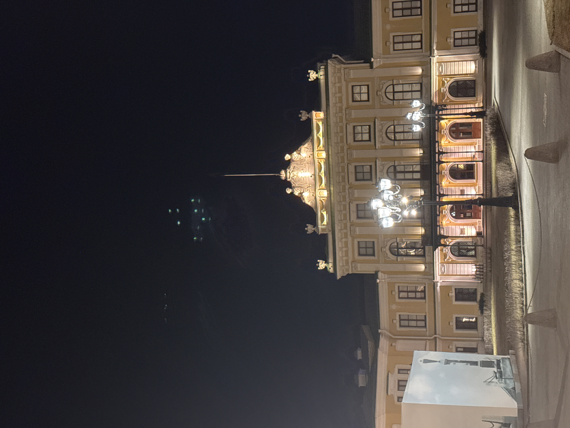 The image shows the Imperial Palace in Tver, Russia, at night. The facade of the palace is illuminated, showcasing its architectural details, including windows, arches, and decorative elements. A street with old-fashioned lamps is visible, and a structure with a modern depiction of the tower is in the foreground. The sky is dark, with a few faint lights visible in the distance.