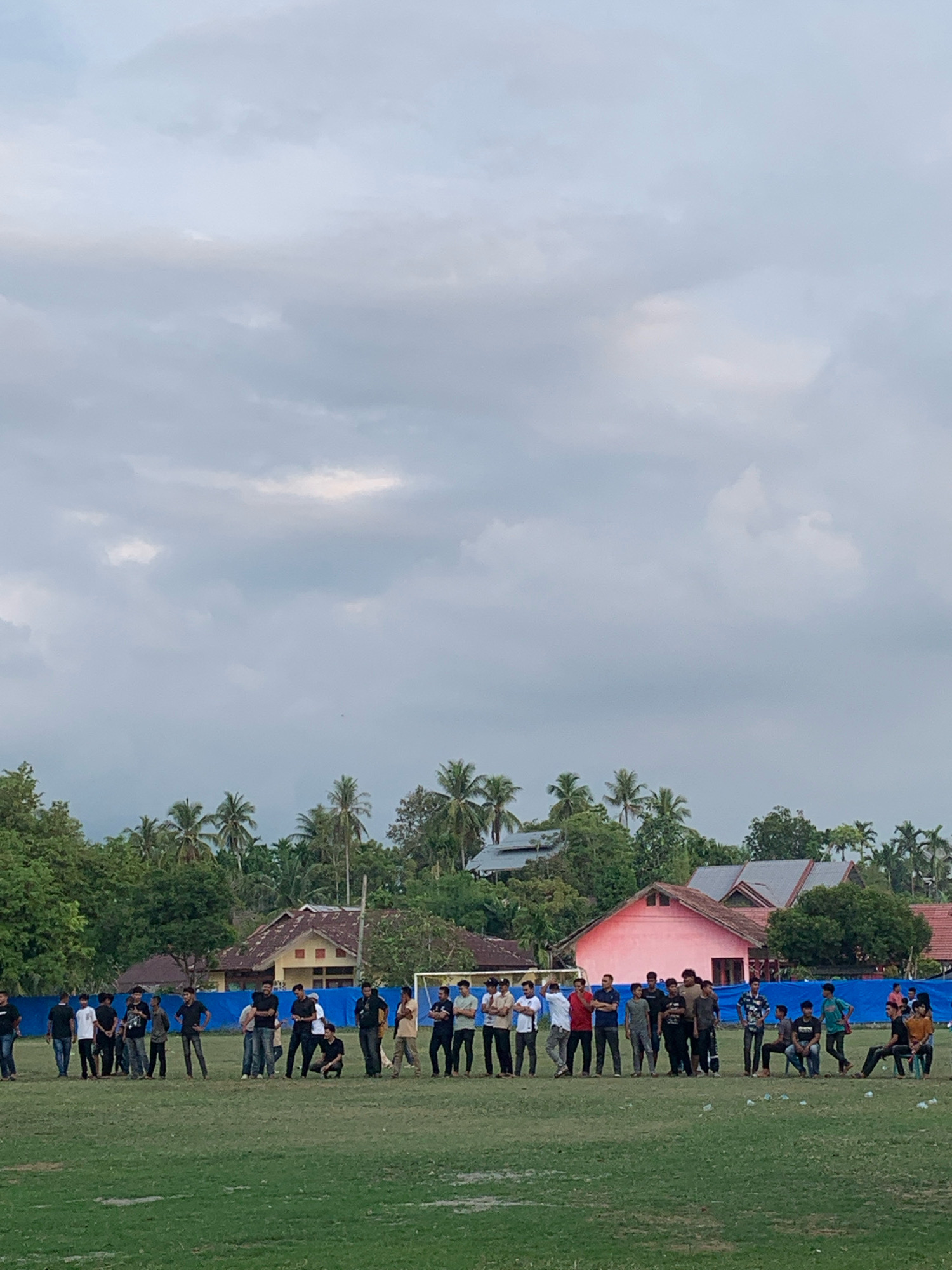 A group of approximately 40 individuals, predominantly young men, are gathered on a grassy field. They are standing and some are sitting. The individuals are dressed in casual clothing, including t-shirts, long-sleeved shirts, and jeans. Some are holding cell phones.
In the background, there are several buildings with red and pink roofs, surrounded by lush green trees, including palm trees. A blue tarp or banner is visible behind some of the people, stretched between poles. A soccer goal is set up on the right side of the field. The sky is overcast with clouds. The overall scene suggests an outdoor gathering or event. The location is the city of Keudelapang, Indonesia.