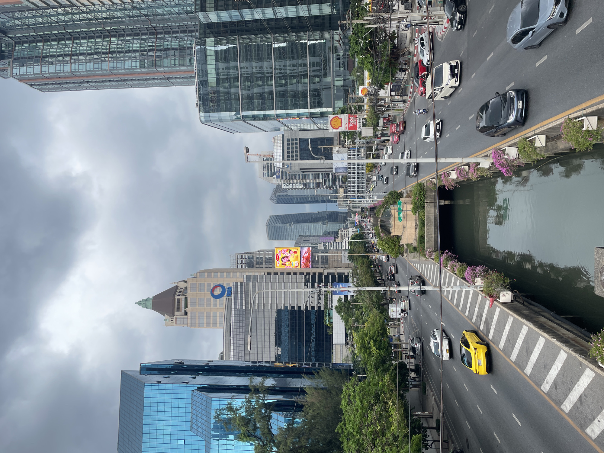 This image shows a busy urban scene in Bangkok, Thailand. The photograph is taken from an elevated perspective overlooking a street lined with modern buildings, including glass-faced skyscrapers and structures with visible corporate branding like a large "O" logo and a Shell gas station sign. There is heavy traffic on the road, consisting of a variety of cars, including sedans, SUVs, and at least one yellow sports car. A canal runs alongside the street, separated by a curb decorated with flowers. The sky is overcast, suggesting a cloudy day. The environment includes dense greenery and trees bordering the street, with billboards and advertisements interspersed among the buildings.