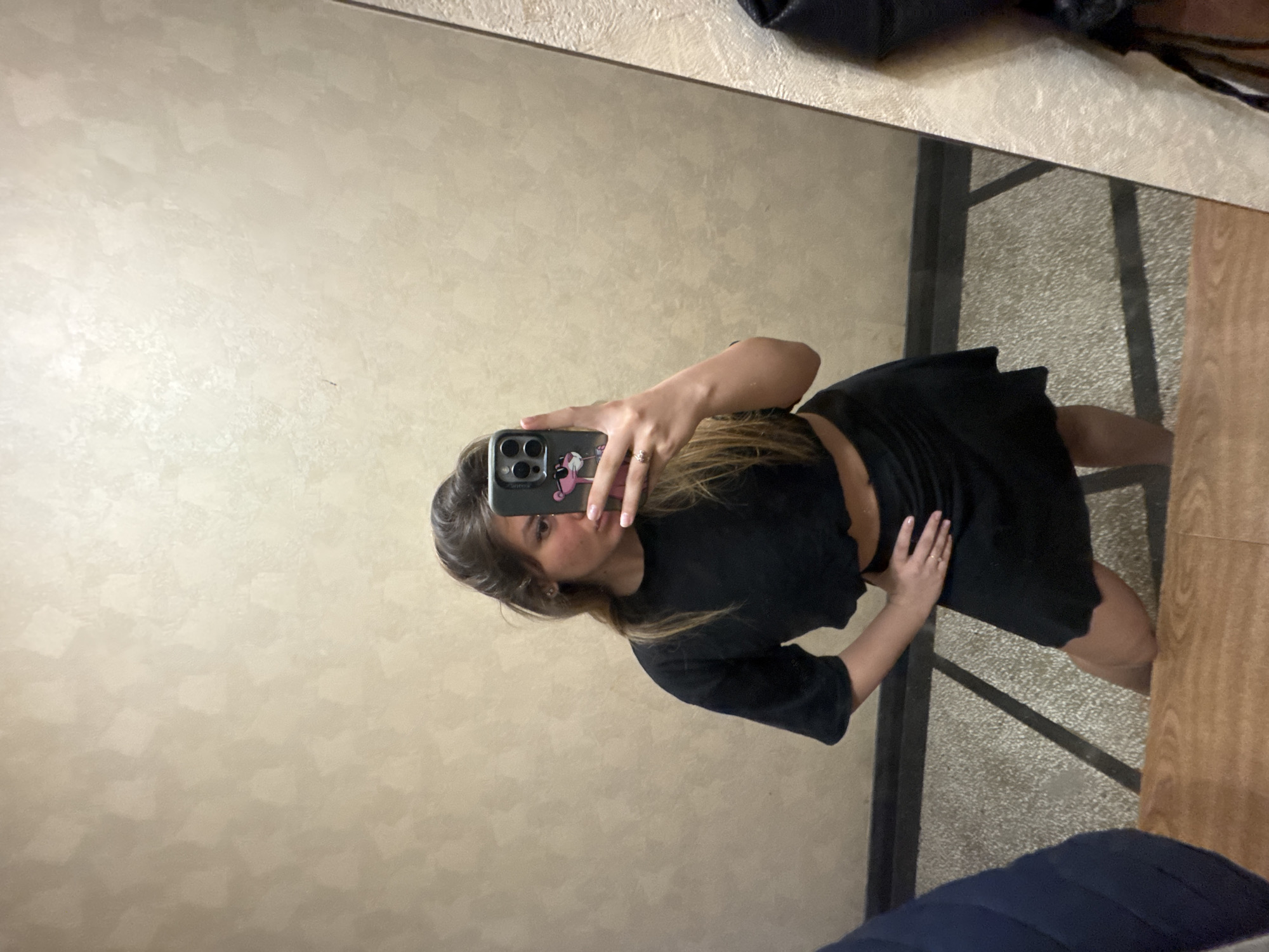 The image is a mirror selfie featuring a young woman in an indoor setting, likely in Sofia, Bulgaria. The original photograph is rotated 90 degrees clockwise.
The subject is a woman with medium-length brown hair, styled with lighter highlights, falling over her shoulders. She is wearing a black, short-sleeved crop top and a matching black skirt or dress. She holds a dark-colored smartphone, possibly an iPhone given the camera array, in her right hand (viewer's left). The phone is encased in a light-colored cover featuring a pink cartoon character design. A ring is visible on her right ring finger. Her left hand (viewer's right) is placed casually on her hip. She maintains a neutral facial expression, looking towards the camera in the reflection.
The reflected background consists of a light beige or tan wall with a subtle, textured pattern. Below the wall, the floor is visible, showing a darker, speckled surface, possibly concrete or a similar material, adjacent to a section of lighter, wood-patterned flooring. A dark, possibly metal, structural element separates these floor types. In the top right corner of the reflection, a dark, indistinct object, potentially clothing or a bag, is seen. Another dark blue fabric object is partially visible in the bottom right corner of the reflected scene. The lighting suggests an indoor environment, with no specific cues about the time of day beyond being well-lit. No discernible text is visible in the image apart from the graphic on the phone case.