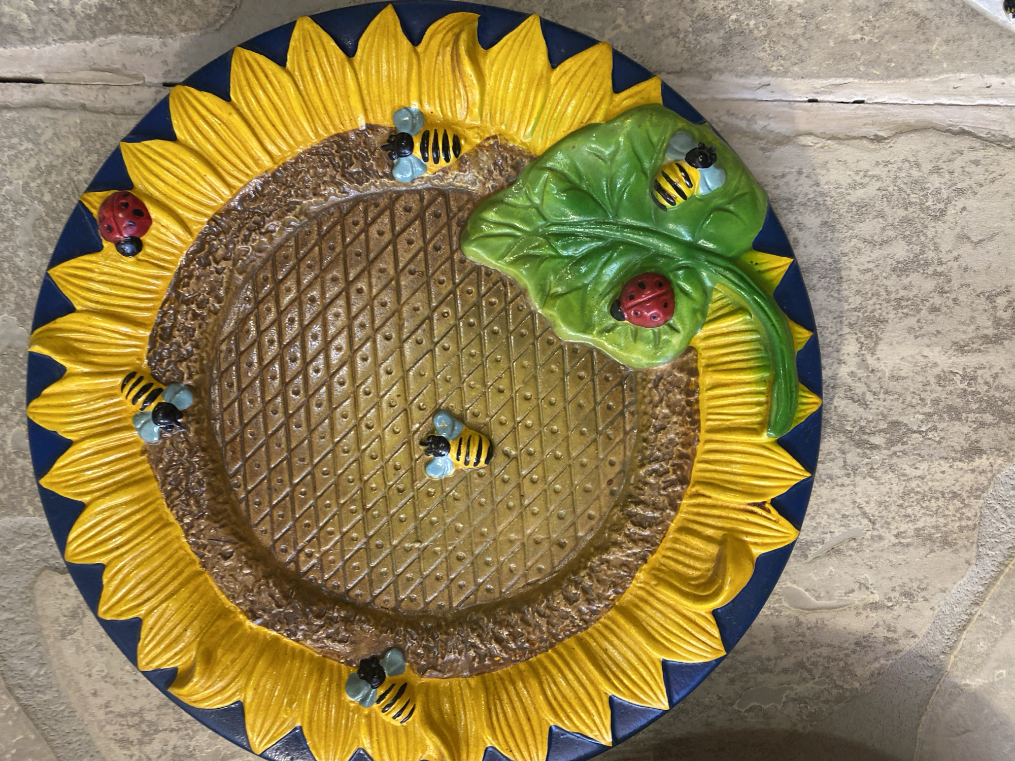 A decorative sunflower plate is displayed outdoors against a stone wall, likely in Novaya Usman', Russia. The plate features a textured brown center resembling a sunflower's seeds, surrounded by bright yellow petals edged in dark blue. Several small, stylized bees with black and yellow stripes and light blue wings are positioned on the plate. Two red ladybugs with black spots are also present, one on a green leaf that sprouts from the side of the sunflower. The plate is mounted on a textured, grey stone wall, with some lighter-colored grout lines visible. The scene is illuminated by natural light.