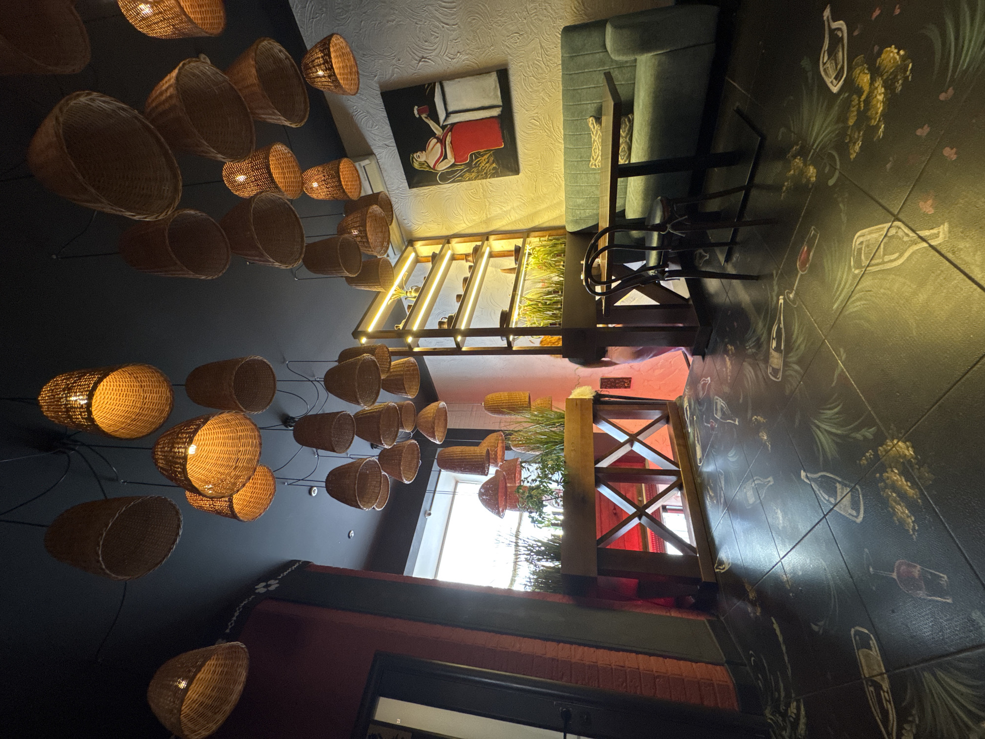 The image depicts the interior of a restaurant in Chernivtsi, Ukraine. The scene is characterized by a dark ceiling adorned with numerous woven basket-shaped pendant lights, casting a warm glow. The walls are a mix of dark red brick on the left and textured white on the right, where a painting of a woman in a red dress holding a drink hangs above a teal sofa.
In the foreground, a table with two chairs sits in front of the sofa. The floor is black and features a decorative pattern of what appear to be wine bottles, glasses, and floral elements, executed in a chalk-like style.
To the left, a wooden structure with red accents and a glass door is visible. Further back, a tall, illuminated shelving unit displays various items, with plants arranged on the lower shelves. The overall ambiance suggests a cozy and somewhat artistic dining establishment. No individuals are clearly visible within the frame, though a figure is partially obscured on the far left.
