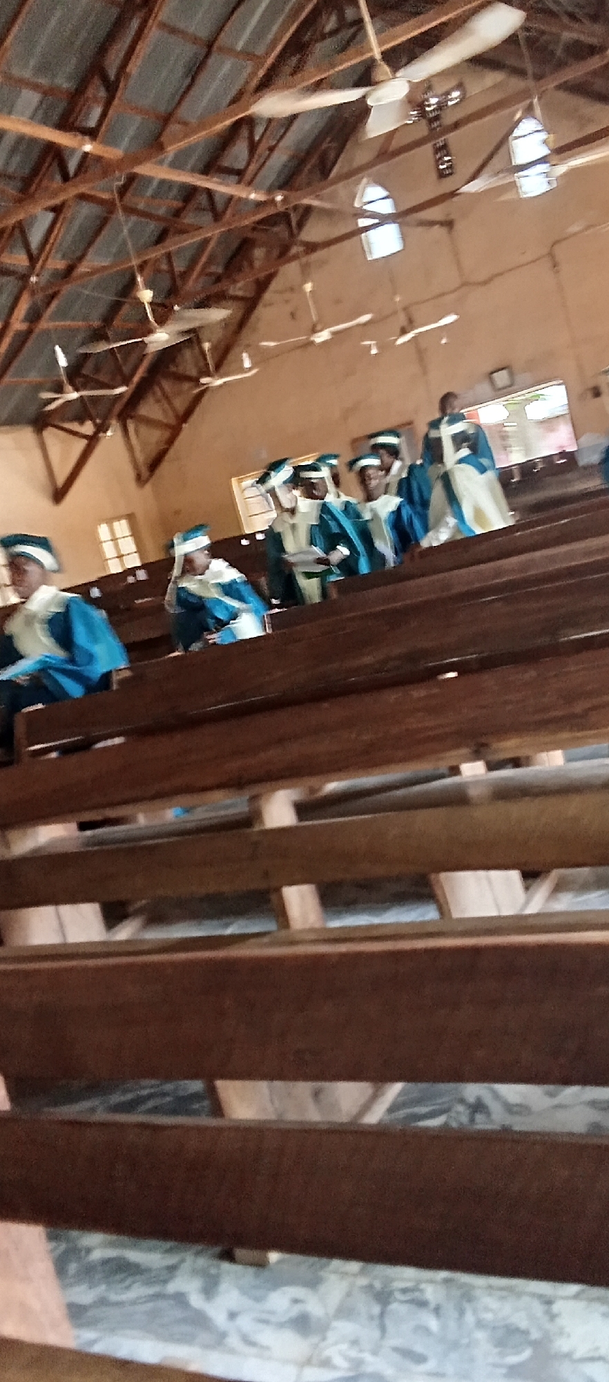 Inside a church-like building in Akwanga, Nigeria, several people wearing blue and white robes are seated on wooden benches. The benches are in rows, one behind the other. The architecture of the building includes a high, open ceiling with exposed wooden beams and several ceiling fans. There are windows near the top of the walls.