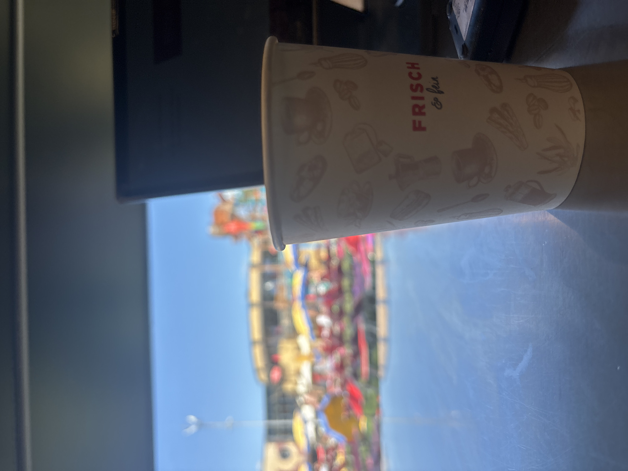 An outdoor scene, possibly a market or festival, is visible in the blurred background. The foreground features a disposable coffee cup with a patterned design and text that reads "FRISCH & fein". The cup is placed on a reflective surface, likely a table or counter, which casts a blueish reflection. A dark object, possibly a card reader or electronic device, is positioned to the right of the cup. The lighting suggests it is daytime. The image does not contain enough information to confirm the location as Gebersdorf, Austria.