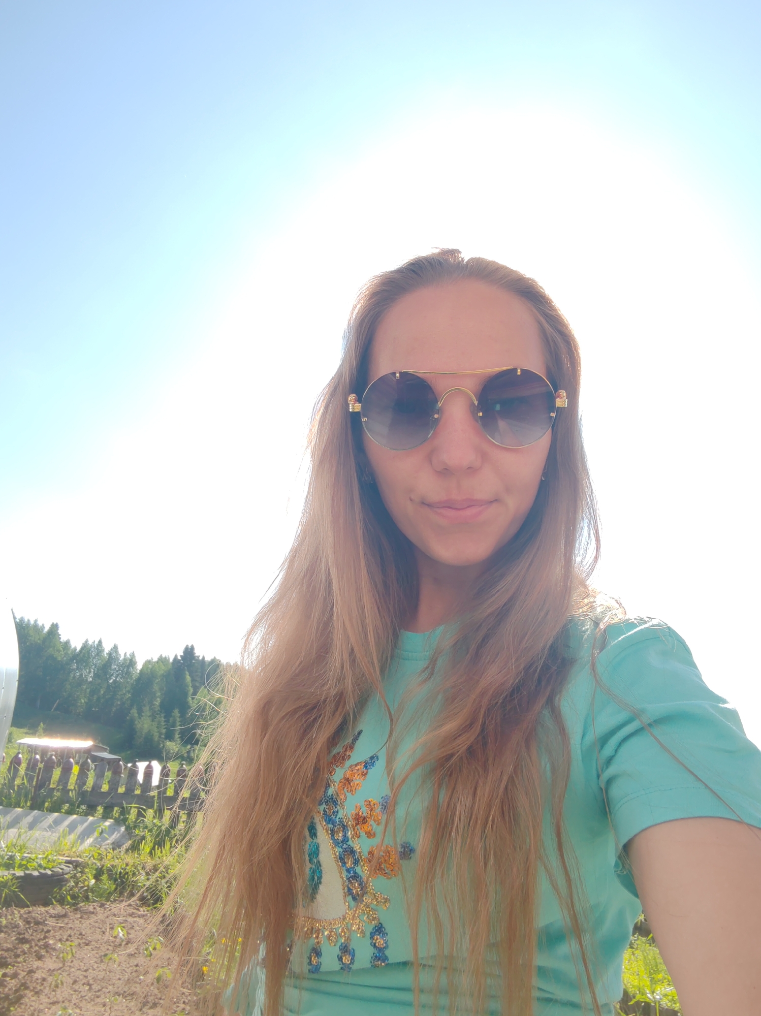 A long-haired woman with light brown hair is taking a selfie outdoors. She is wearing a teal t-shirt with a floral design and large, round, gold-rimmed sunglasses. The background is a bright, sunny sky with some trees and a wooden fence visible in the distance. The ground appears to be dirt and grass. The location is identified as Chukhlem, Russia.