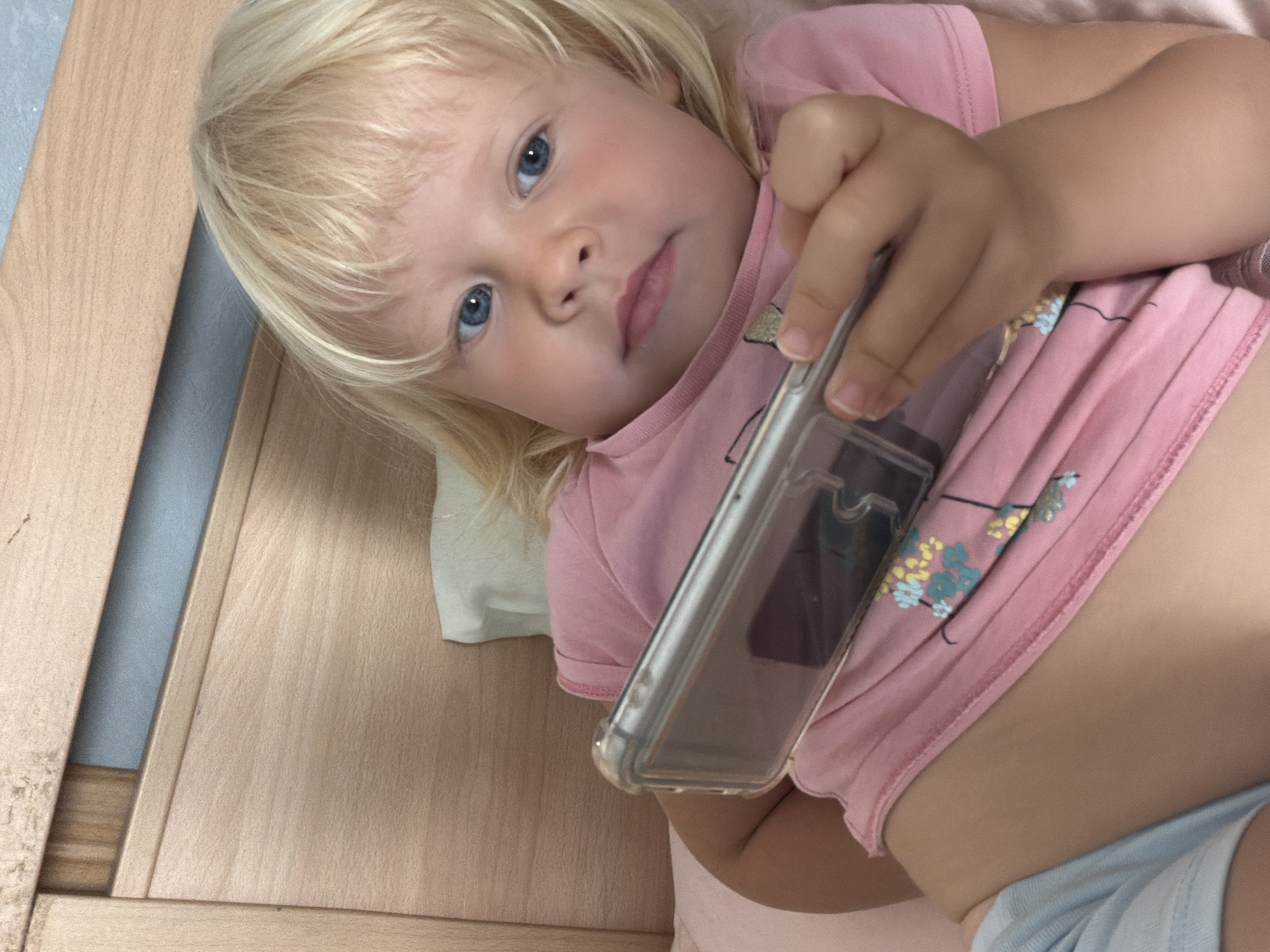 A young child with blonde hair and blue eyes is lying in bed, holding a smartphone. The child is wearing a pink t-shirt with a floral pattern and light blue shorts. The bed has a wooden headboard and is covered with pink and white bedding. The overall environment suggests an indoor residential setting, possibly a bedroom. There is no visible indication of the location being in Sochi, Russia.