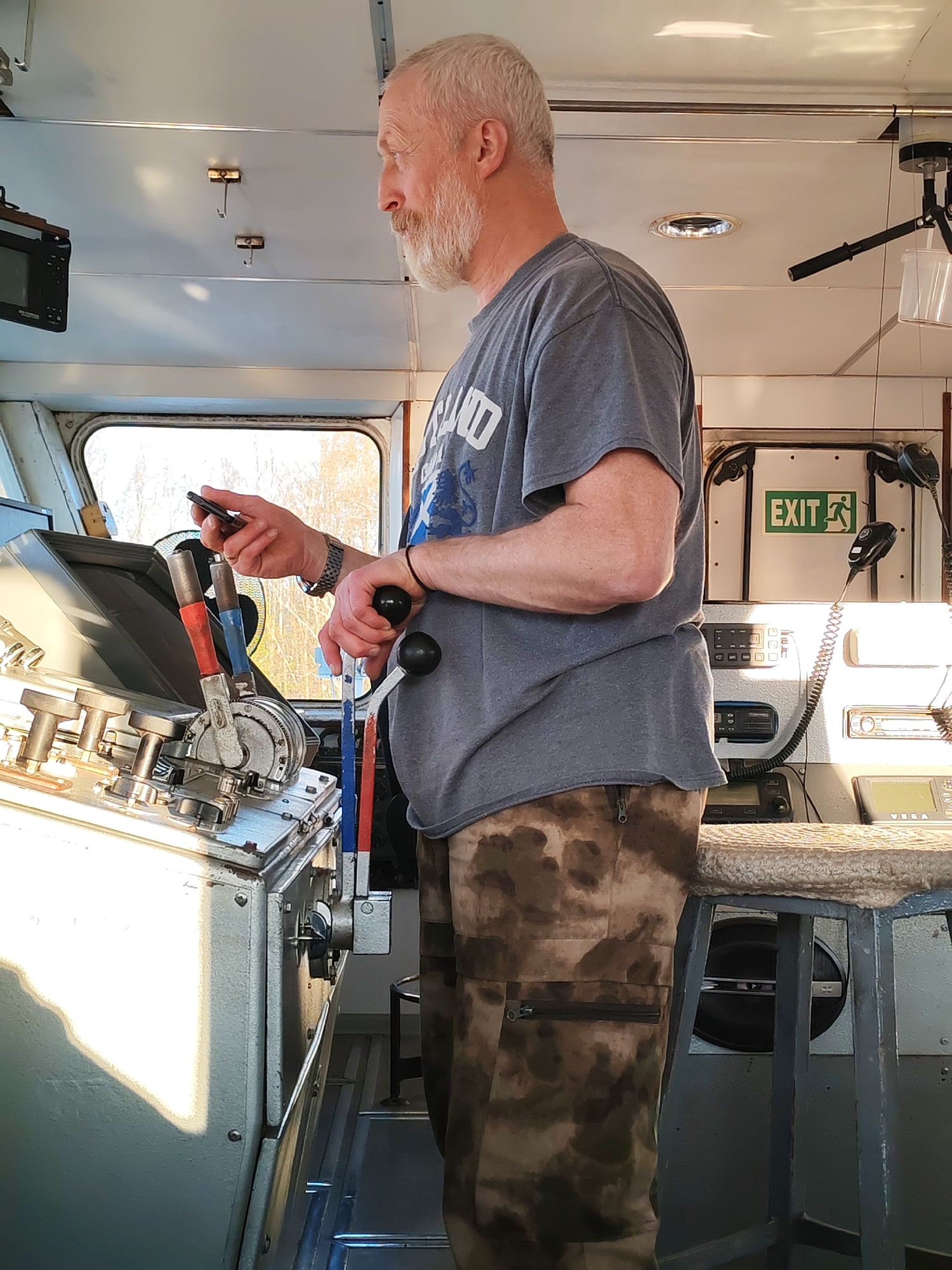 The image depicts an older man with gray hair and a beard standing inside what appears to be the control cabin of a train or boat. He is wearing a gray T-shirt and camouflage pants. He is holding a smartphone in his left hand and operating levers with his right hand. The controls are situated on a metallic console with various levers and gauges. Behind him, there is an "EXIT" sign and what appears to be radio communication equipment. There is a stool in the background. A window is visible, showing an exterior scene. The man's attention is focused on the instruments and the mobile phone. It is unknown whether the location is in Kurdiug, Russia.