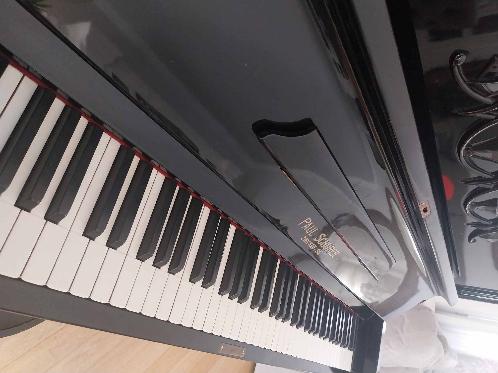 A close-up, angled shot shows a black upright piano. The piano lid is open, revealing a row of white and black keys. The brand name "PAUL SCHOBER" is visible in gold lettering on the piano's surface, along with "ZWICKAU, SA." The polished black surface of the piano reflects ambient light and the surrounding environment, including a blurred glimpse of a beige sofa and a window with sheer curtains in the background. The lighting suggests an indoor setting, possibly a living room. The image is taken from a low angle, emphasizing the keys and the glossy finish of the instrument.
