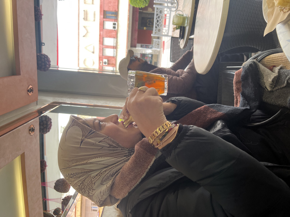 This image depicts an indoor cafe scene, identified as being in Meknes, Morocco, due to the visible "CAMEL" signage on a red building observed through the window, likely referencing Café Camel. In the foreground, a woman is captured in profile, facing right. She wears a light-patterned headscarf, a textured scarf in shades of rust and dark blue/grey, and a black puffy jacket. Her left wrist is adorned with multiple gold-toned bracelets, including one prominent chain-link style and several thinner ones with small colored elements. She is actively biting into a light-colored food item held in her left hand, while her right hand holds a clear glass mug filled with an amber liquid containing mint leaves, presumably tea.
Behind her, to the right and further back, a man is partially visible, wearing a light-colored baseball cap and dark sunglasses, seated at a separate table. The cafe interior features light-colored round tables and dark, possibly wicker, chairs. Overhead, the ceiling displays reddish-brown panels with recessed lighting and decorative clusters of items resembling dried fruits suspended. Through the large window, an urban street scene is visible, including a distinct red-orange building with large white letters spelling "CAMEL," a white ornamental metal fence, and green foliage. On a table in the background, another clear mug containing a green liquid is present. A white bowl with a yellow food item is partially visible in the bottom right corner.