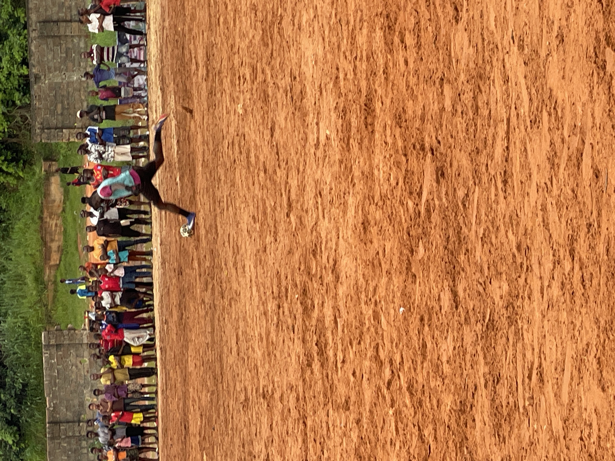 In an outdoor, dirt-field setting, a person in athletic attire is captured in mid-kick, striking a soccer ball. The individual wears a teal and pink jersey, black shorts, and blue athletic shoes. The background features a crowd of spectators, some of whom are identifiable as men, women, and children, dressed in casual clothing. Beyond the spectators, a low wall made of concrete blocks is visible, with lush green foliage extending upwards. The overall scene depicts a lively game of soccer taking place in an informal urban or rural environment.