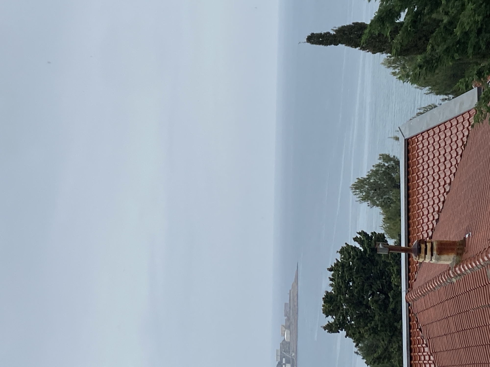 The image shows a view of the Adriatic Sea from what appears to be a residential area in Šušanj, Montenegro. The sky is overcast and the sea is calm with a light haze. A terracotta-tiled roof of a building dominates the right side of the image, with a chimney extending upwards. In the background, a few trees are seen. In the distance over the sea is what appears to be a man made structure.
