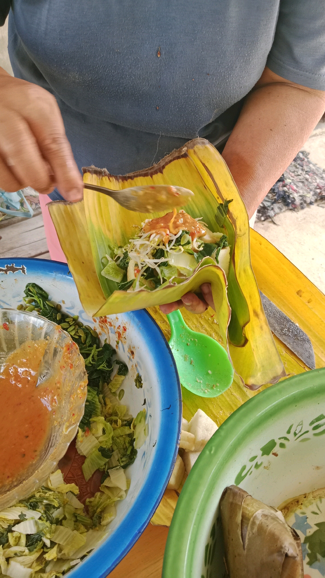 A person is preparing a dish wrapped in a banana leaf. The dish contains vegetables and is being topped with a peanut sauce. The person is holding a spoon and adding the sauce to the vegetables. There are other bowls of vegetables and sauce nearby. The scene appears to be outdoors, possibly in a rural area of Bulukerto, Indonesia. The background shows a rough, textured surface.