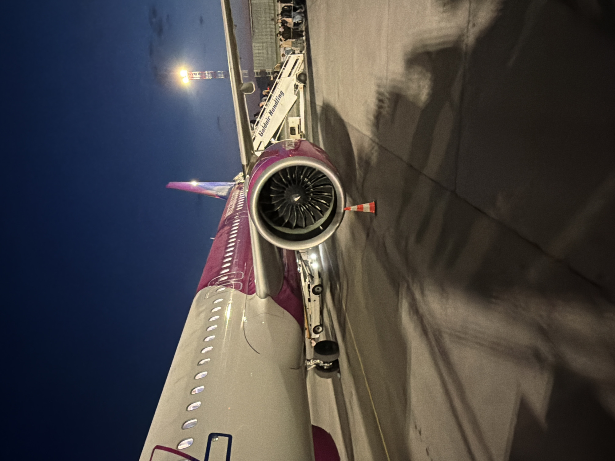 The image shows an airplane at an airport at night. The plane's engine is visible, along with part of the plane's tail. A set of stairs is attached to the plane with people walking on it. The words "Goldair Handling" are visible on the stairs. The location is implied to be Sofia, Bulgaria.