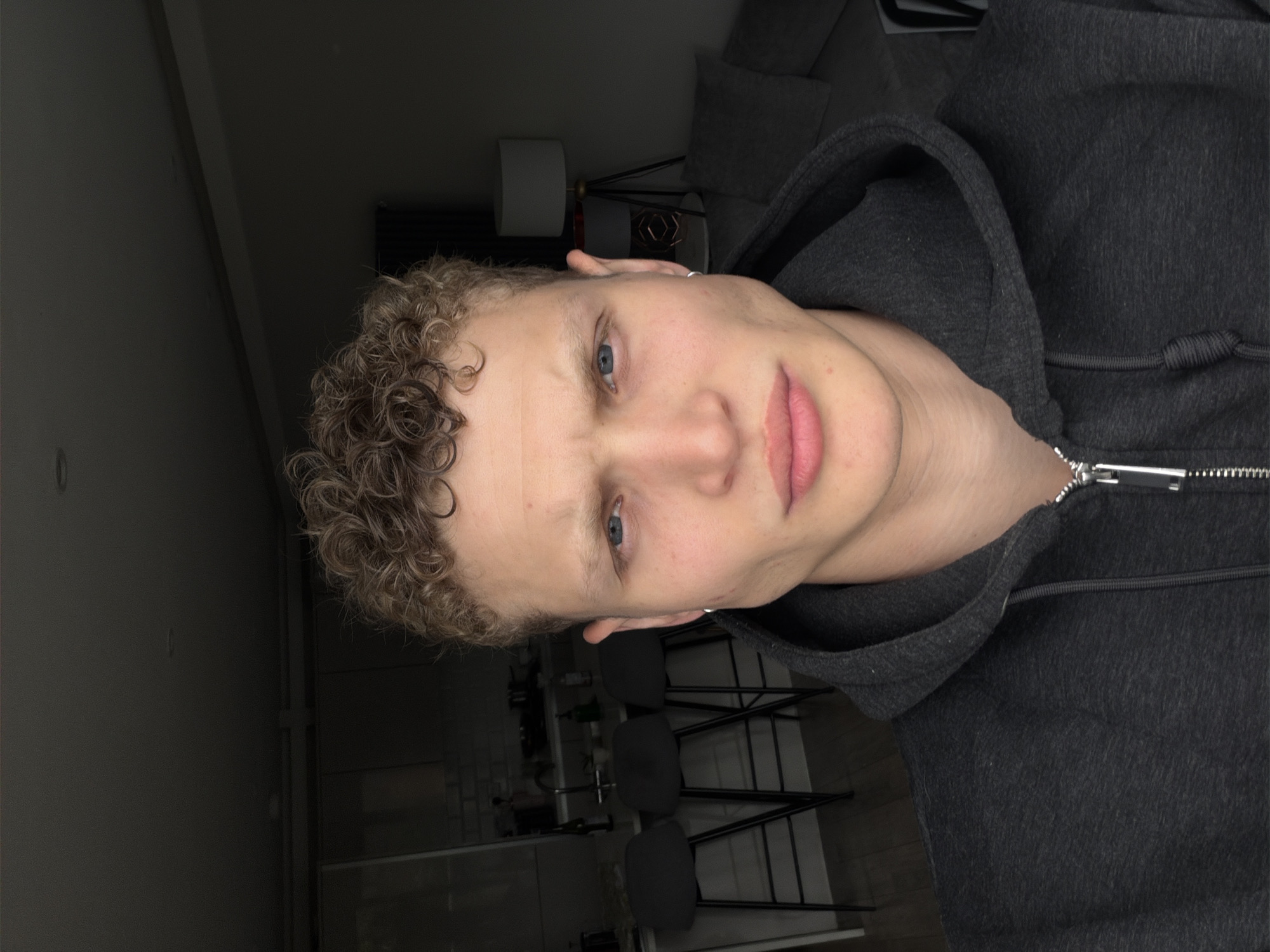 A young adult male with light brown curly hair and blue eyes looks directly at the camera, his eyebrows slightly furrowed and lips pressed together in a serious expression. He wears a dark grey hooded sweatshirt with a silver zipper. A small silver hoop earring is visible in his right ear. The scene is an interior located in the city of London, United Kingdom, specifically an open-plan apartment or studio. To the viewer's left, a kitchen area is visible with a light-colored counter, three dark bar stools with black metal frames, and a tiled backsplash. Several bottles and a faucet are on the counter. To the viewer's right, a living area contains a dark grey sofa with a light grey cushion and a tripod floor lamp featuring a drum shade and a geometric base. The overall lighting is dim. A dark wooden floor is visible on the left.
