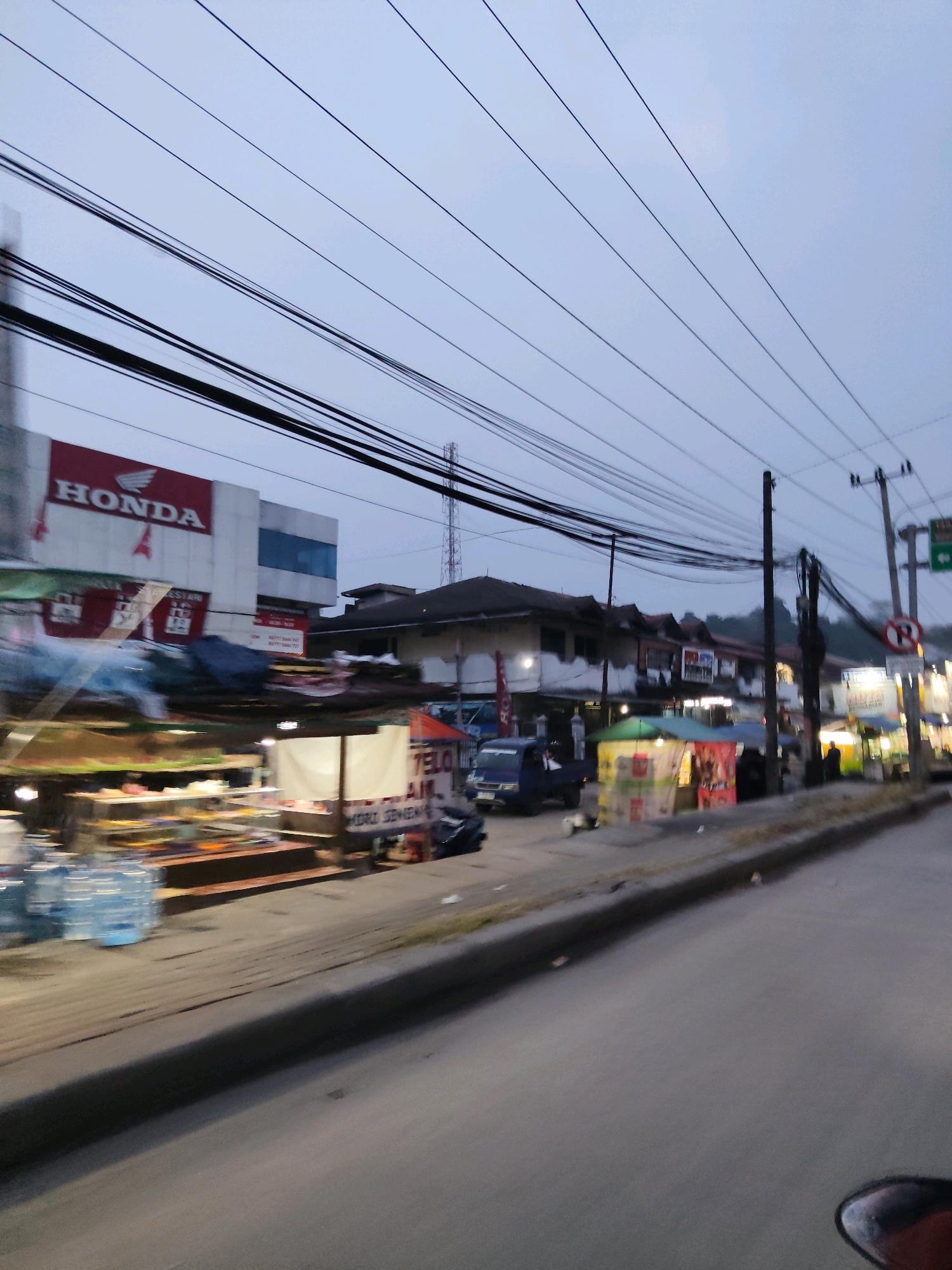 The image depicts a street scene in Kaduagung Tengah, Indonesia, likely during early morning or dusk, given the muted lighting. The foreground shows a paved road with a curb. Beyond the curb, a sidewalk area features several stalls with awnings and displays of goods, including what appears to be food items on shelves and stacked water bottles. A blue utility vehicle is parked near one of the stalls.
In the background, a Honda dealership is visible, characterized by a prominent red and white sign. Adjacent to it are multi-story buildings with windows and balconies. Further down the street, more stalls and businesses are present, some illuminated by artificial lights. Overhead, a complex network of electrical wires stretches across the frame, supported by utility poles. In the distance, a telecommunications tower can be seen. The overall environment suggests a commercial area with roadside vendors. No individuals are clearly identifiable or engaged in specific actions.