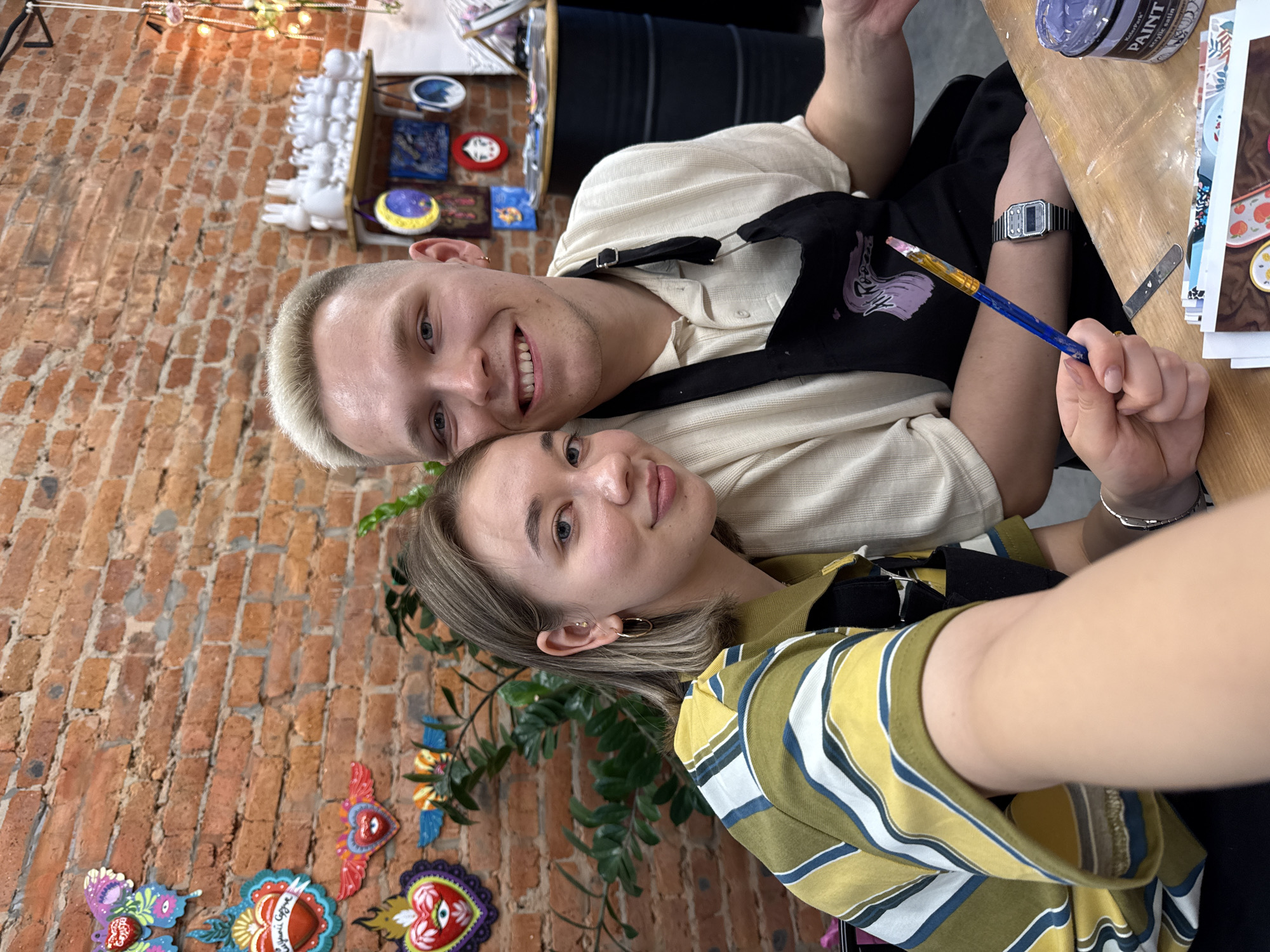 A selfie photograph captures two individuals indoors, likely in an art studio or creative space.
The person at the top of the frame has short, bleached blonde hair and is smiling. They are wearing a light-colored, button-down shirt and a black apron. A digital watch is visible on their left wrist. They are holding a paintbrush.
Below them, the second individual is also smiling. They have short, light brown hair. They are wearing a yellow and green striped shirt, with a black apron visible underneath.
The background includes a brick wall adorned with colorful, decorative heart-shaped art pieces. Shelves display painted objects and figurines. A can of paint is visible on a table.
The photo's orientation is off-kilter, slightly tilted. The location is identified as the city of Chaikovsky, Russia.