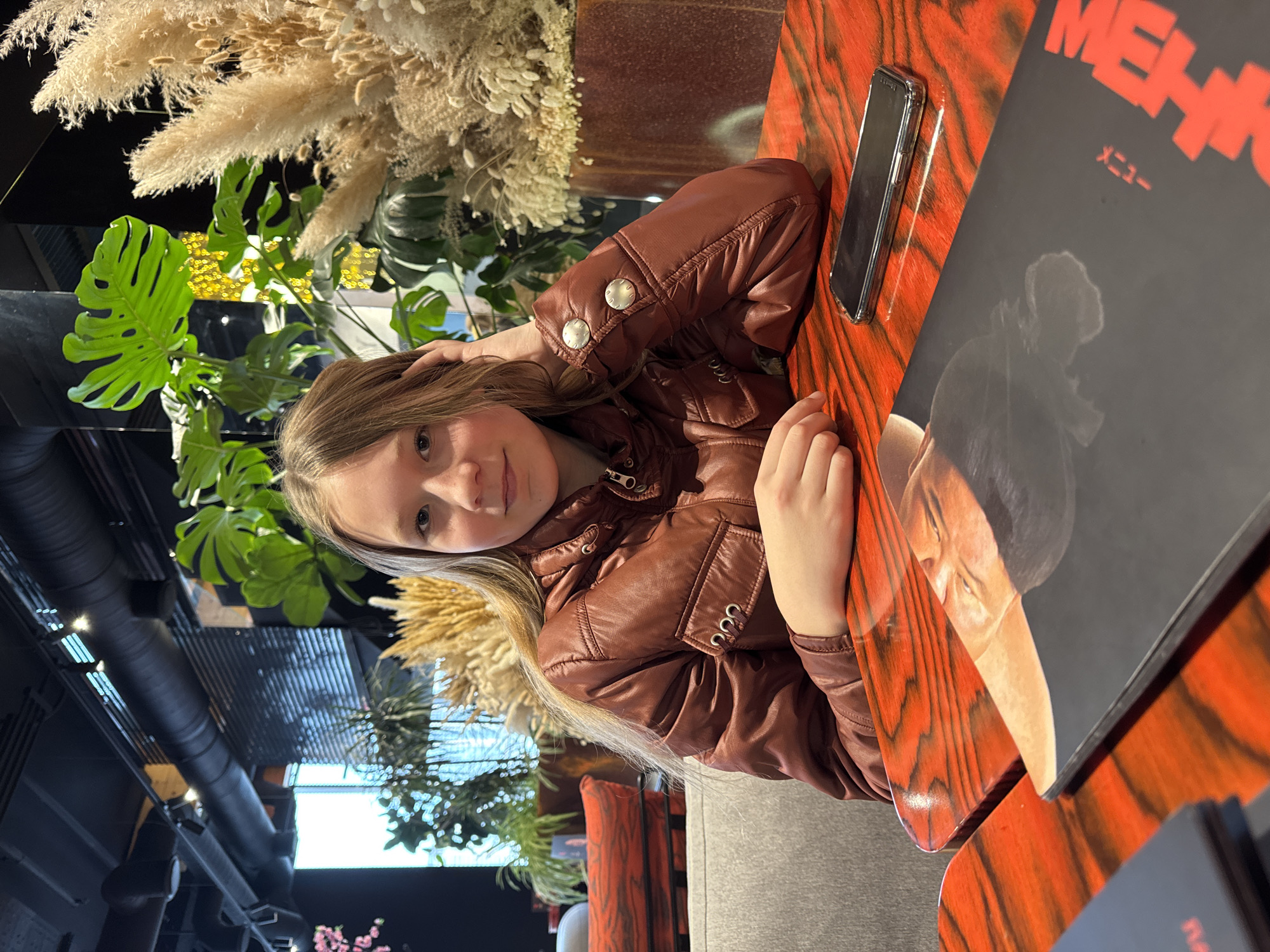 A young girl sits at a red wood-grain table at a restaurant in Rostov-on-Don, Russia, visible on the menu in English and Japanese. She wears a brown leather jacket and rests her head on her hand, looking at the camera. A phone and a menu are on the table. Behind her are plants and window views of the city. The ceiling has exposed pipes and metal grates.