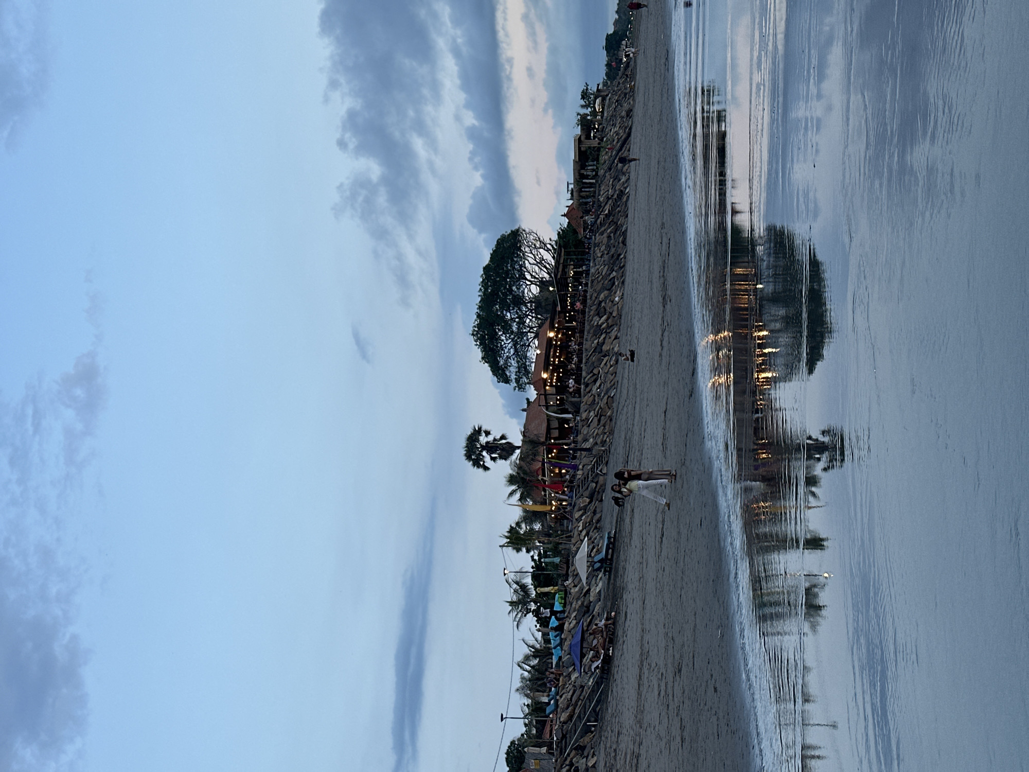 The image depicts a beach in North Kuta, Indonesia, at dusk. The sky is cloudy, with the clouds reflected in the wet sand near the water. Along the beach are numerous people. Some are sitting on beach chairs under umbrellas, while others are walking along the shoreline. There's a restaurant with string lights hanging from a large tree. The lights are also reflected in the water. Palm trees are scattered along the beach. A seawall comprised of rocks separates the sand from the land beyond.