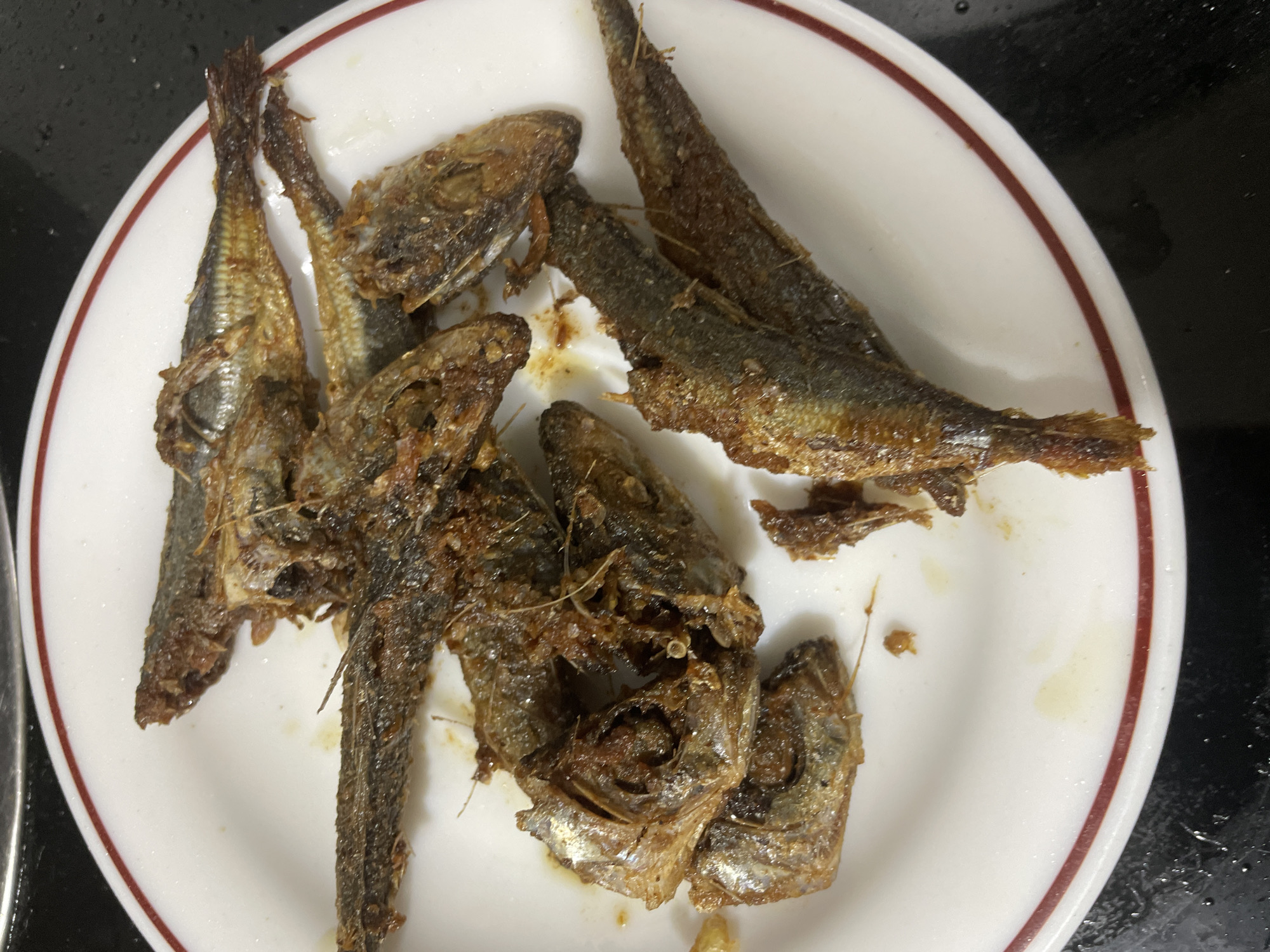 A white plate with a red trim sits on a dark, reflective surface. The plate contains several small, fried fish. The fish are arranged in a pile, with some overlapping. They appear to be whole, with heads and tails visible, and are a golden brown color from being fried. Some of the fish are broken or partially consumed, revealing their flaky flesh. There are small droplets of oil and some scattered remnants on the plate. The lighting suggests an indoor setting, possibly a kitchen or dining area. The background is dark and slightly blurred, with some subtle reflections. The overall impression is of a simple, prepared meal. There is no visible text in the image. The context provided indicates the location is Davao City, Philippines.