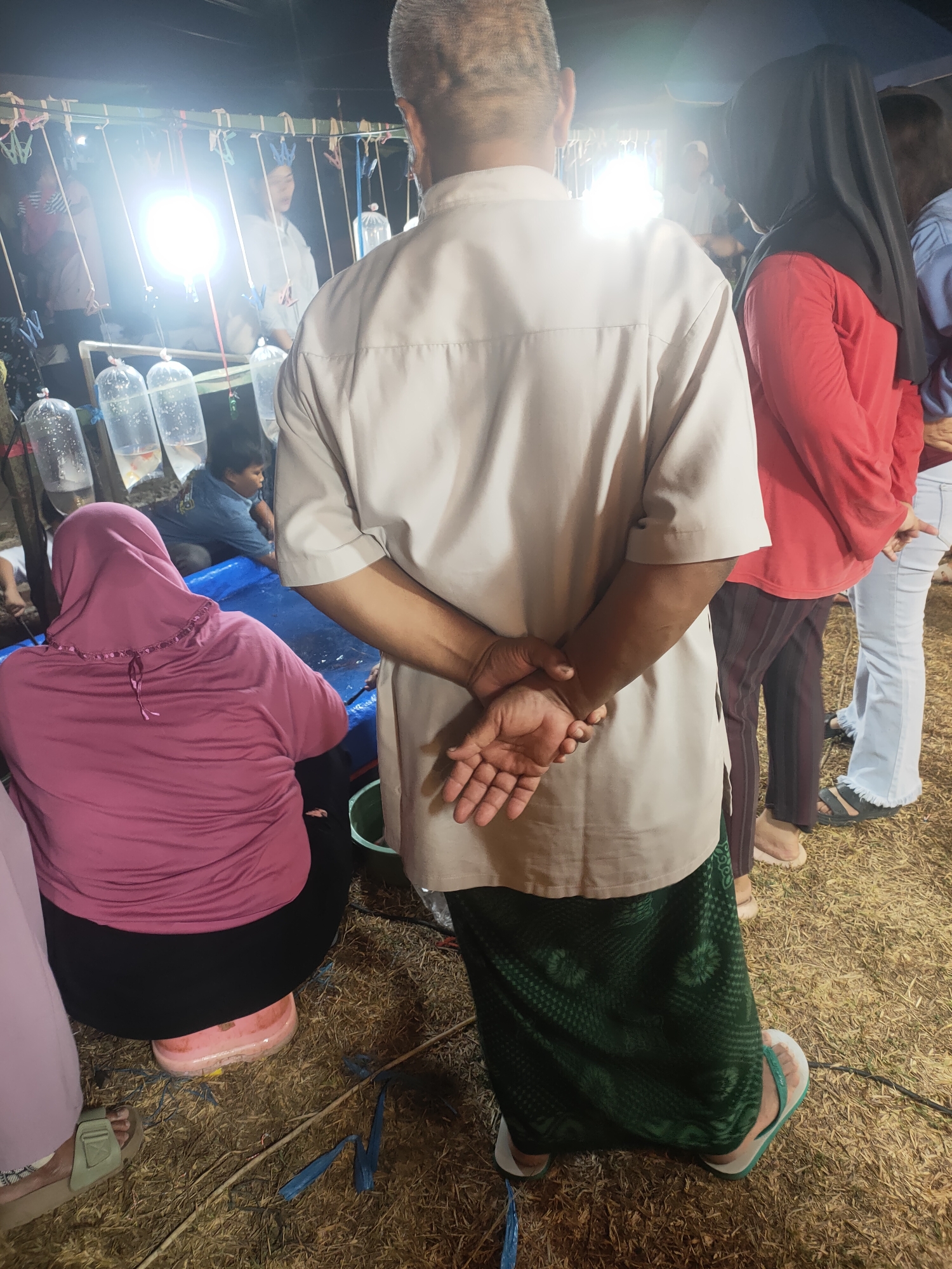 A group of people are gathered at an outdoor night market in Subang, Indonesia. The foreground is dominated by the backs of two individuals. A man in a light-colored shirt and a green sarong stands with his hands clasped behind his back. To his left, a woman in a pink hijab and a black skirt is seated. Behind them, a display of fish in plastic bags is illuminated by bright lights. Other individuals, including women in hijabs, are visible further back in the scene, suggesting a busy market atmosphere. The ground is covered with dry grass and straw.
