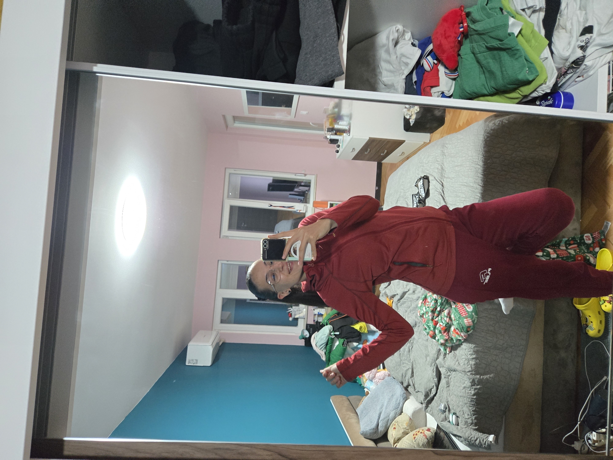 The image shows a person in red sweats and a matching top taking a selfie in a mirror. The reflection reveals a room with light pink walls, windows, and blue paint on the wall below the windows. There's a bed with a grey comforter visible. Above the mirror, clothes are stacked in a closet. Some yellow shoes are on the bed. The location in Pleven, Bulgaria, cannot be ascertained from the image.