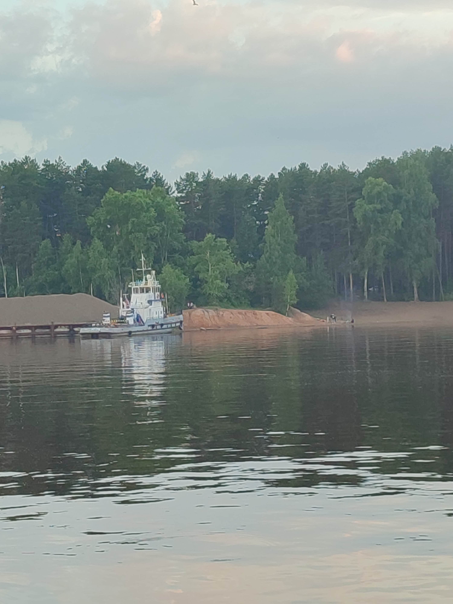 An outdoor, medium shot captures a body of water in the foreground, with ripples distorting the reflections of a tree-lined shore in the background. The sky above is overcast with soft, pink and grey clouds.
On the water, a white tugboat is docked next to a barge laden with sand. The tugboat has a white cabin with blue accents and a white flag at its stern.
The shore is a sandy beach bordering a dense forest of green trees. In the distance, on the beach, a small group of people are visible, with smoke rising from what appears to be a campfire. The overall atmosphere suggests a peaceful, natural setting, possibly during a late afternoon or early evening.