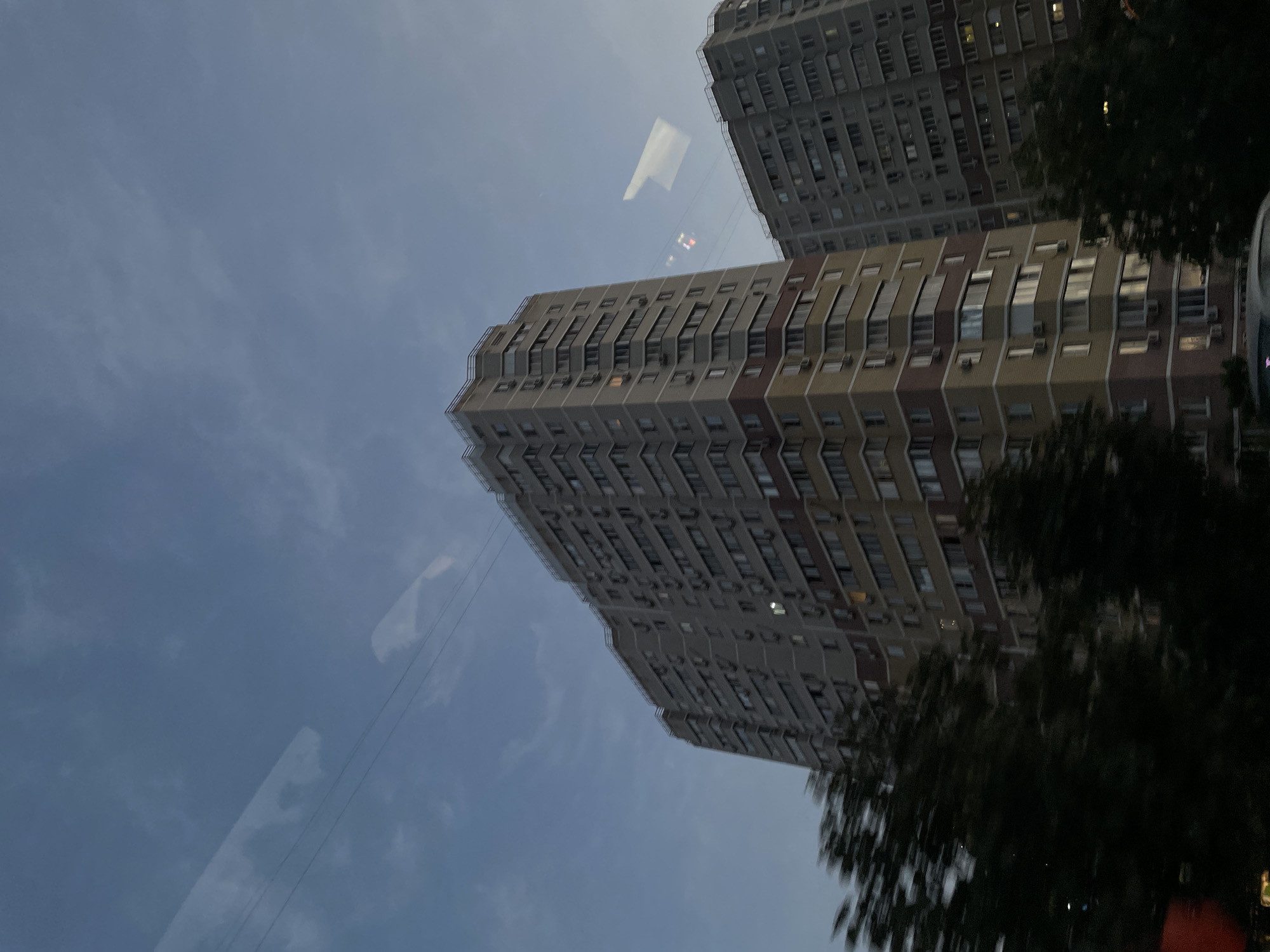 The image shows two tall, multi-story residential buildings in Rostov-on-Don, Russia, against a twilight sky with scattered clouds. The building in the foreground is taller and has a segmented facade with alternating bands of light gray, yellow, and maroon. It features numerous windows and air conditioning units. The building in the background is slightly shorter and appears to be a uniform light gray color, also with many windows and AC units. Dark green trees are visible in the foreground, partially obscuring the lower sections of the buildings and appearing somewhat blurred, suggesting movement. There are reflections on what appears to be a glass surface in front of the viewer, showing abstract shapes and light, possibly from streetlights or vehicle lights. The overall scene depicts an urban residential environment during the evening.