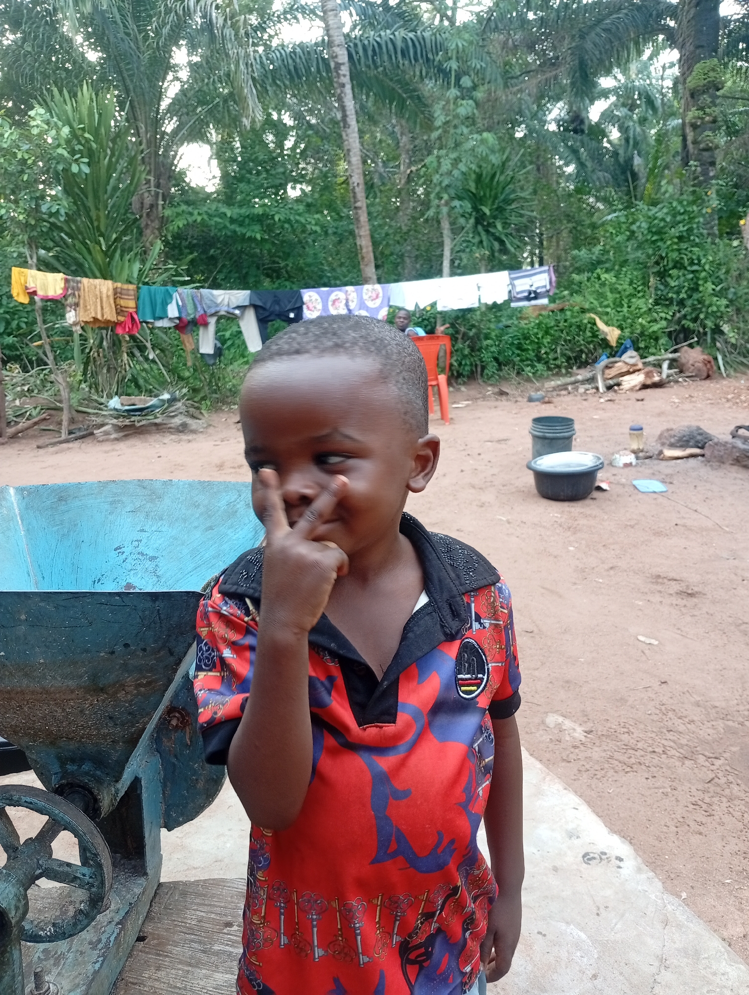 A young boy with dark skin and short hair, wearing a red and blue patterned t-shirt, stands in front of a blue metal structure in a dirt yard. His left hand is raised, with two fingers extended in a peace sign. In the background, laundry hangs on a line between two poles, and lush green vegetation with palm trees is visible. The scene appears to be outdoors in a rural or semi-rural setting, likely in Adda, Nigeria. Several buckets and some debris are scattered on the ground.
