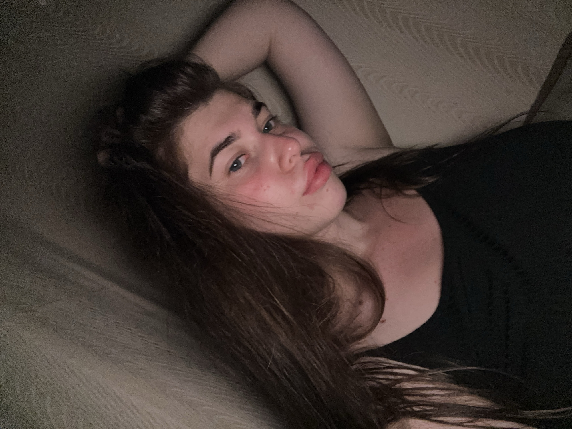 A young woman with long brown hair is lying down, her head resting on her arm, which is bent at the elbow and placed behind her head. She is wearing a black tank top. Her face is turned towards the camera, and she has a slight blush on her cheeks. The background appears to be a textured wall. The lighting is dim, casting shadows on the scene. The image was captured indoors. The location is stated to be in the city of Мещёры, Russia.
