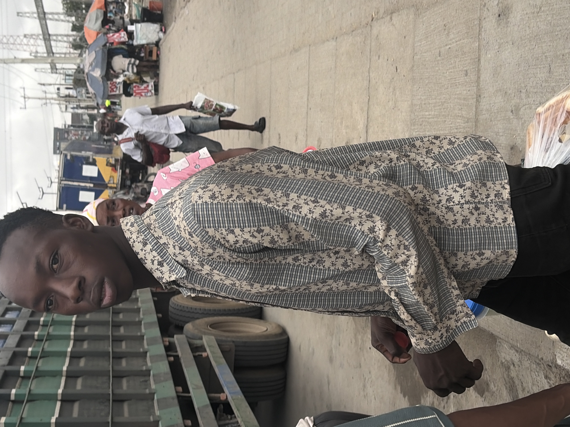 In Igboefon, Nigeria, a young Black man stands in the foreground, his body angled to the left, wearing a patterned shirt and black pants. He is looking towards the viewer. Behind him, a large green truck is parked. To the right, other people are visible, including a woman in a pink shirt and a man in a white shirt carrying a bag. The background shows a bustling street scene with market stalls and overhead power lines. The ground is paved with concrete. A large clear plastic bag containing items is on the ground in the lower right corner.