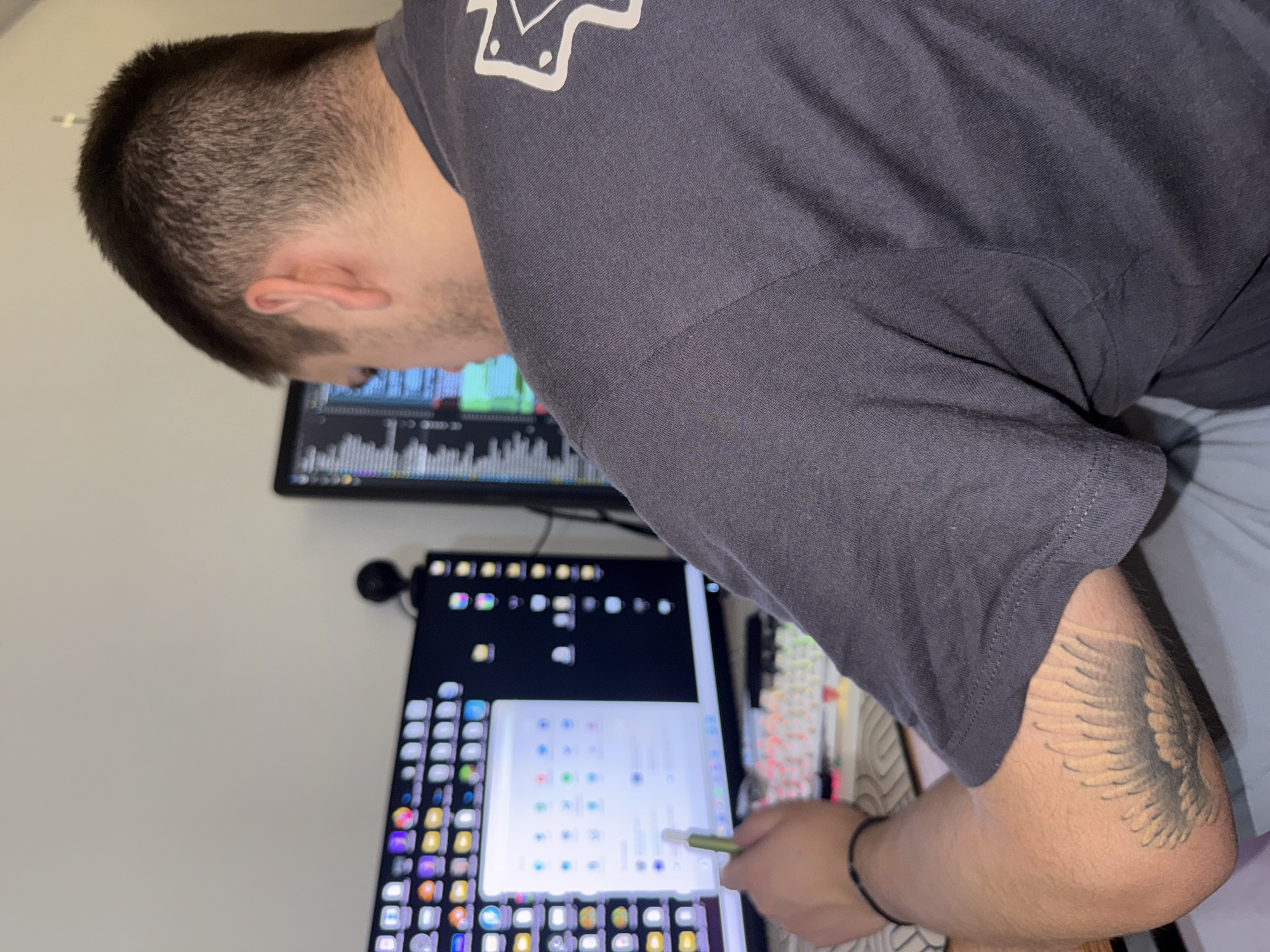 A medium shot depicts a man hunched over a desk, likely indoors. He has short brown hair and is wearing a dark gray t-shirt with a white logo on the upper left. The man is looking at multiple computer screens, one of which is a tablet with a stylus in his hand. The screen displays a grid of application icons. Another monitor behind the tablet appears to be displaying data charts. There is a mechanical keyboard visible on the desk. The man has a tattoo on his right arm. The background is a plain white wall. There is no information suggesting the photo was taken in Plovdiv, Bulgaria.