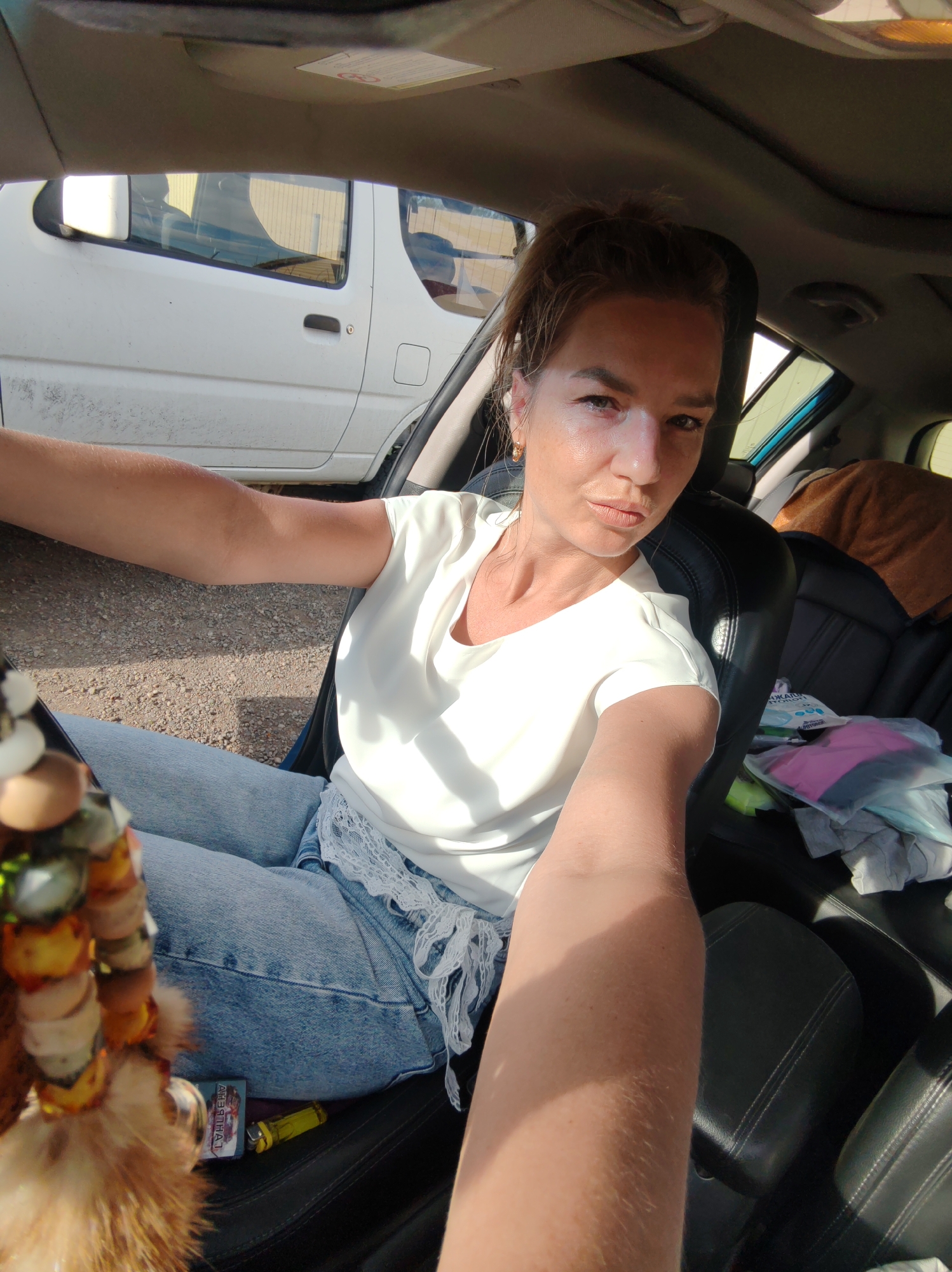 The image shows a woman in the driver's seat of a car. She is wearing a white shirt and blue jeans. Her hair is pulled up in a messy bun, and she is wearing gold hoop earrings. The car appears to be parked on a gravel road, and there is another white vehicle parked behind her. The interior of the car is visible, including black leather seats and a sun visor. There are some bags and a blanket on the back seat. A keychain with feathers and beads hangs from the rearview mirror. A pack of cigarettes and a yellow lighter are visible on the floor of the car. The lighting suggests it is daytime, and the sun is casting shadows. The location is the city of Paratunka, Russia.