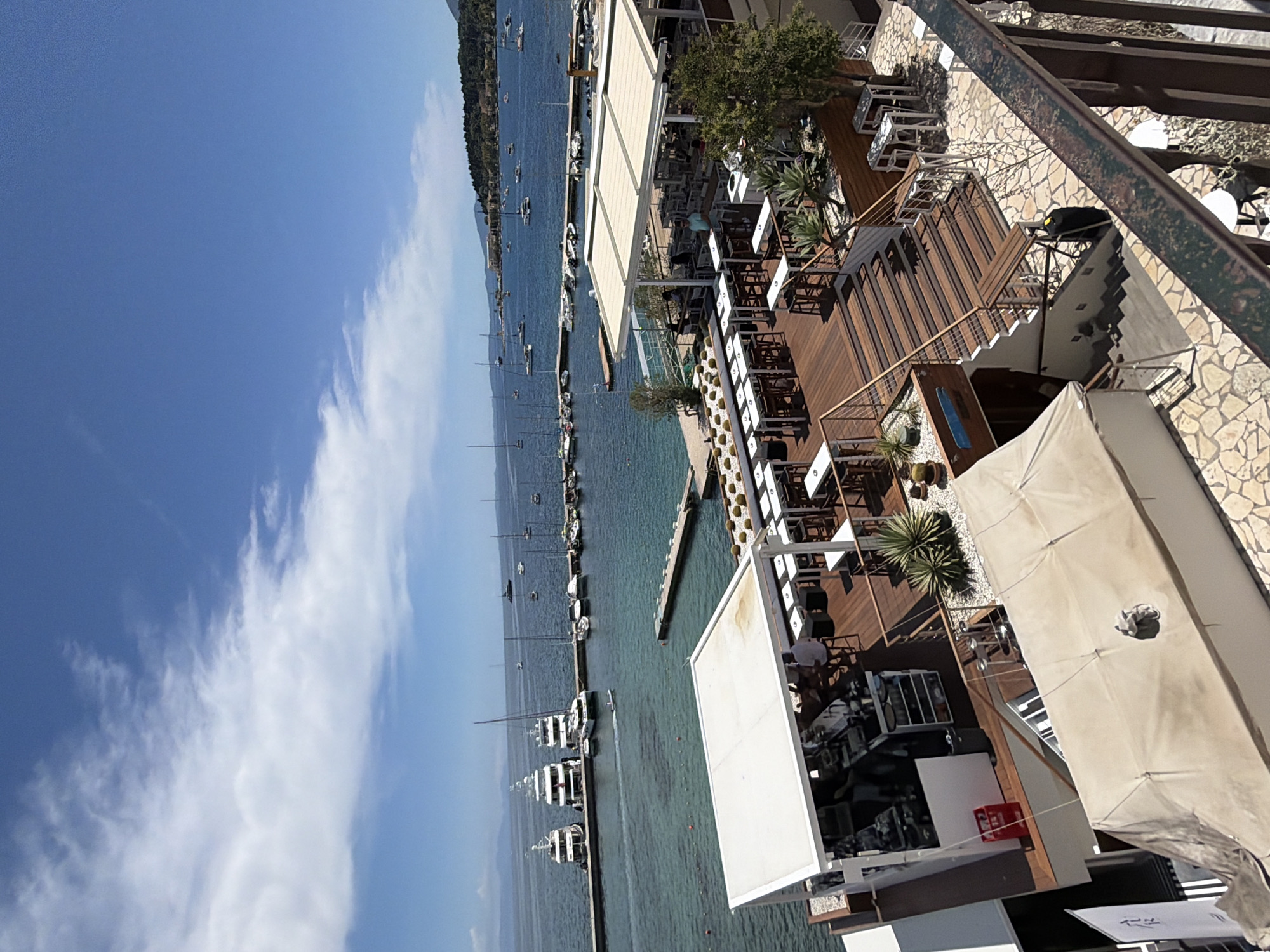This image captures a vibrant coastal scene, likely a resort or restaurant complex, overlooking a bay in Corfu, Greece. The photo is taken from an elevated perspective, possibly a balcony.
The upper portion of the image is dominated by a clear, bright blue sky with scattered, wispy white clouds, indicating sunny weather. Below, a calm, turquoise-blue sea stretches out, filled with numerous boats. Many sailboats with tall masts are anchored or sailing in the distance, while several motor yachts are docked along a pier that extends into the water. The far coastline is visible, featuring rolling hills covered in lush greenery.
On the right side of the frame, a multi-level outdoor dining or lounge area is prominently featured. This stylish establishment boasts extensive wooden decking and stone-paved pathways. It is furnished with numerous white tables and chairs, some shaded by large, off-white umbrellas and pergolas, suggesting an inviting al-fresco setting. Planters containing small trees and succulents are interspersed throughout the terraces, enhancing the aesthetic. A few people can be seen on different levels of the terrace, appearing to be dining or relaxing.
Notable details include strong sunlight, indicating midday or early afternoon, and the distinct shadows cast by the structures. The overall atmosphere suggests leisure and vacation. Visible text includes "FIRE" on a red object, possibly a fire extinguisher or sign, on one of the lower terraces, and "W-FI" (likely "Wi-Fi") on a white board. A metal railing is visible in the immediate foreground on the right, emphasizing the high vantage point of the shot.