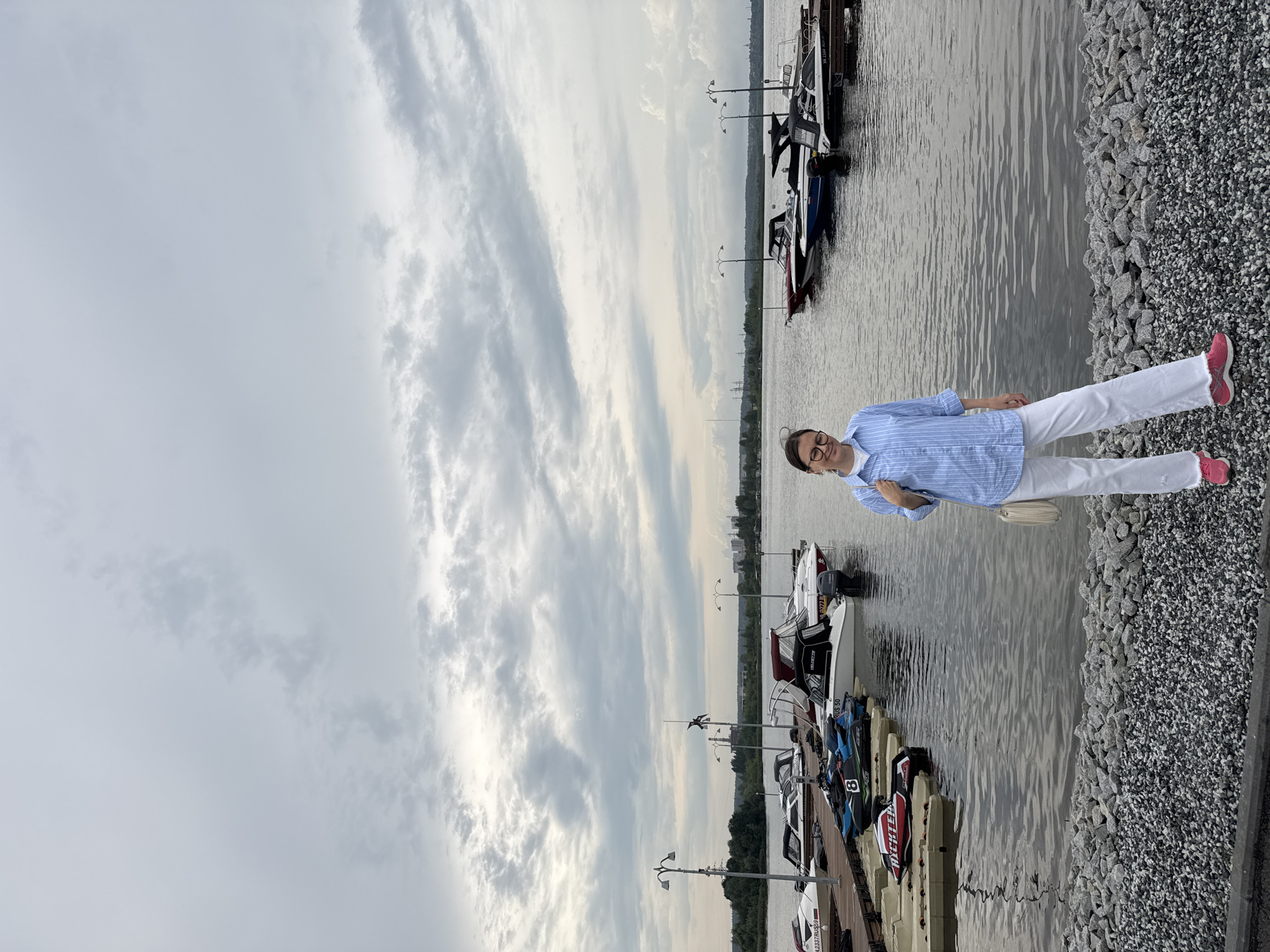A woman with glasses and a light blue striped shirt and white pants stands on a pebble shore in front of a marina. Several boats, including jet skis and speedboats, are docked in the water. The sky is overcast with dramatic clouds. The location is the Marina Club "Zolotaya Rybka" (Golden Fish) on the Izh River in Izhevsk, Russia.