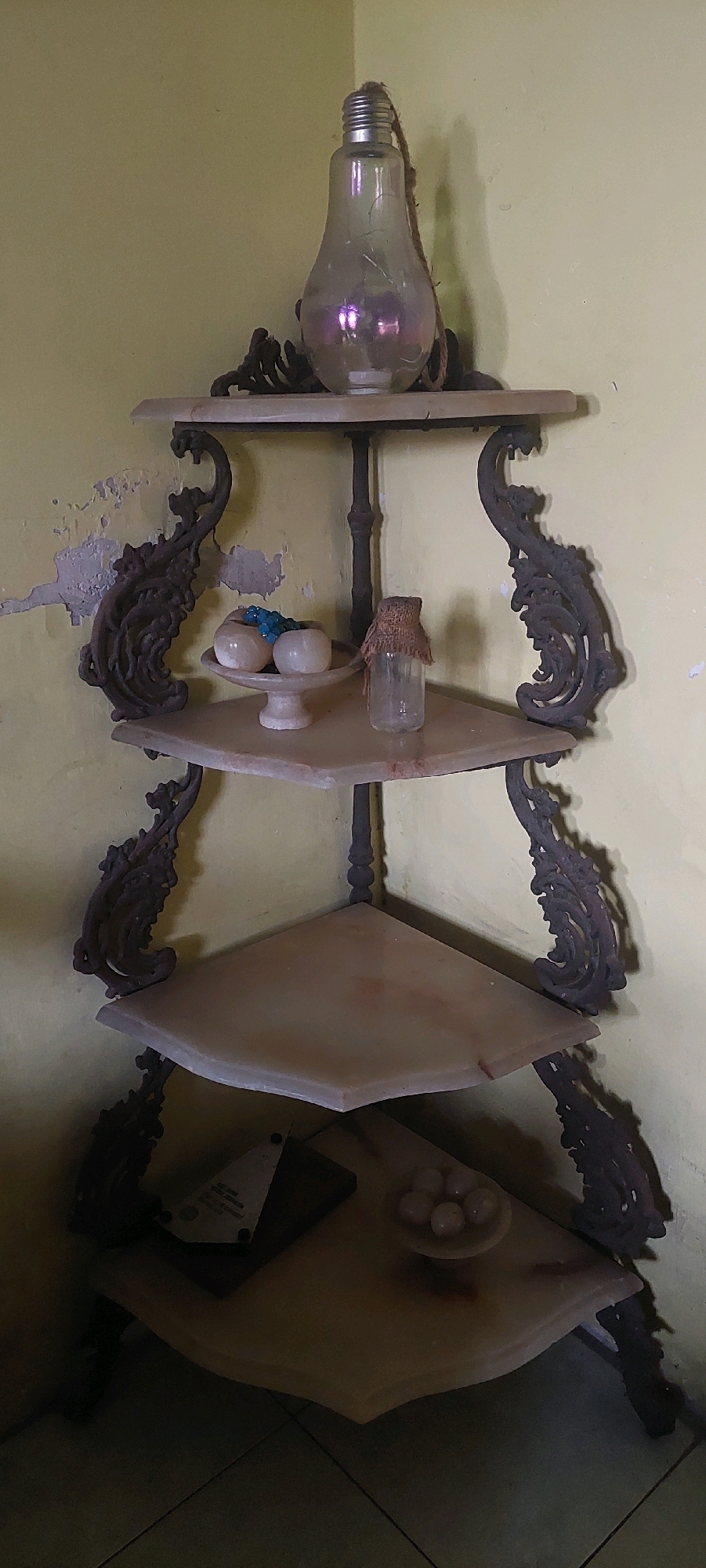 An antique, four-tiered corner shelf unit stands against a pale yellow wall in a room. The shelf unit is made of dark metal with decorative scrolled accents and features marble or stone shelves.
On the top shelf, a large, clear glass light bulb with a thick rope hanging from its metal cap is placed centrally. The bulb exhibits a pearlescent, iridescent sheen.
The second shelf holds a small, light-colored pedestal dish containing several round, pale objects, possibly decorative balls or stones, and a small cluster of bright blue beaded objects. To the right of the dish is a small, clear glass bottle covered with a textured, brown material secured by twine.
The third shelf is bare.
The bottom shelf contains two objects. To the left, a dark, flat object with a triangular, reflective surface, possibly a plaque or award, is visible. To the right, a small pedestal dish holds several pale, round objects, similar to those on the second shelf.
The wall behind the shelf unit shows signs of wear, with peeling paint in patches. The flooring appears to be tiled. The scene is dimly lit, with shadows cast by the shelf unit and its decorative elements. No people or animals are present. The location is unknown.