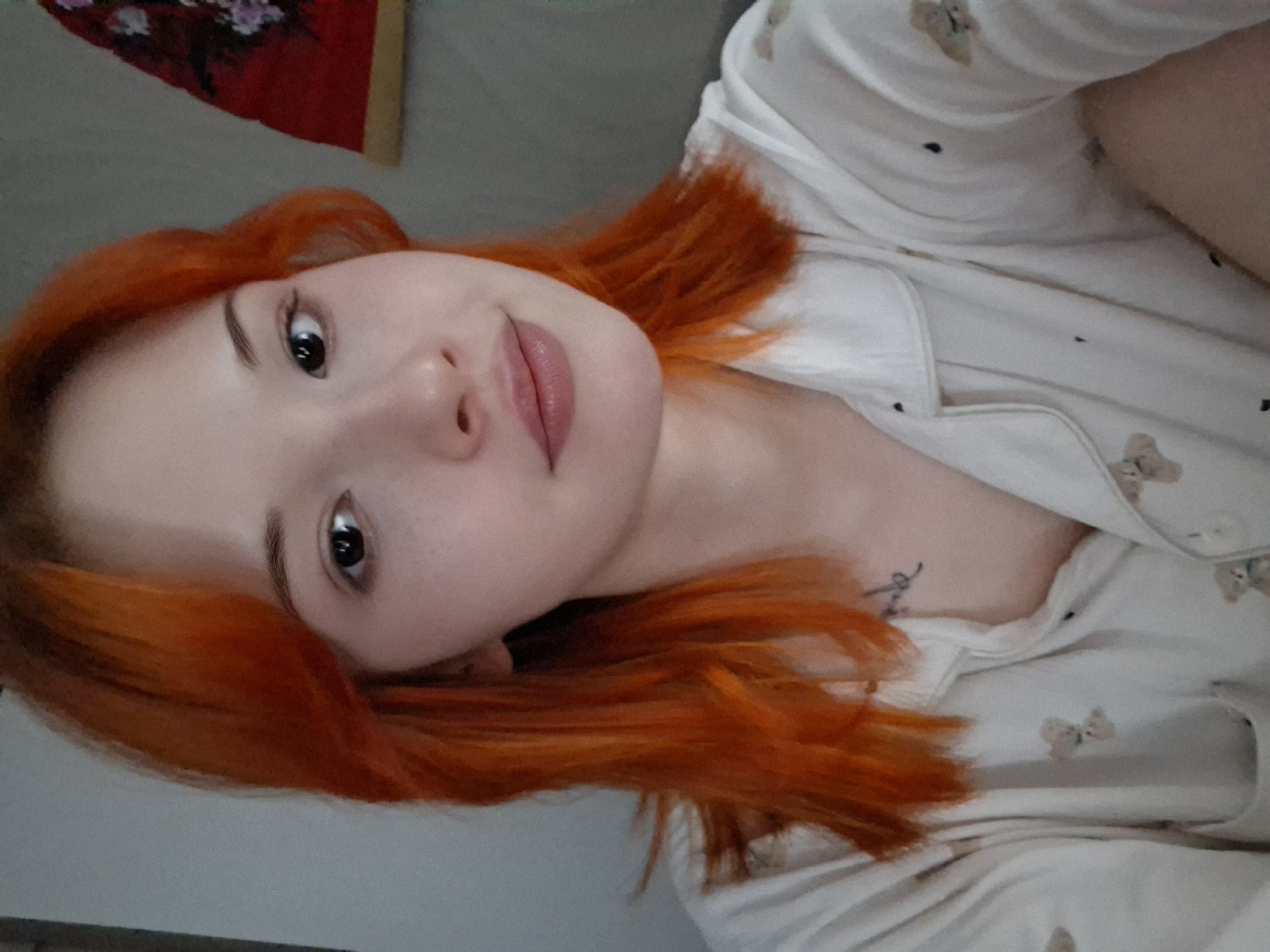 The image is a close-up, slightly rotated selfie of a young woman with striking, vibrant reddish-orange hair. She has fair skin, dark eyes, and well-defined eyebrows, looking directly at the camera with a gentle, soft smile. Her lips are a natural pink color. She is wearing a light-colored, button-up top, likely pajamas or loungewear, adorned with a repeating pattern of small, light brown teddy bears and tiny dark dots. A dark, cursive-style tattoo is visible on her upper chest, near her collarbone, partially obscured by her hair and clothing, though the text itself is not clearly legible. In the upper left background, a red object with floral or intricate patterns is partially visible, suggesting an indoor setting. The lighting is soft and diffused, contributing to a relaxed and intimate atmosphere. The woman appears calm and content. This photograph was taken in Chaikovsky, Russia.