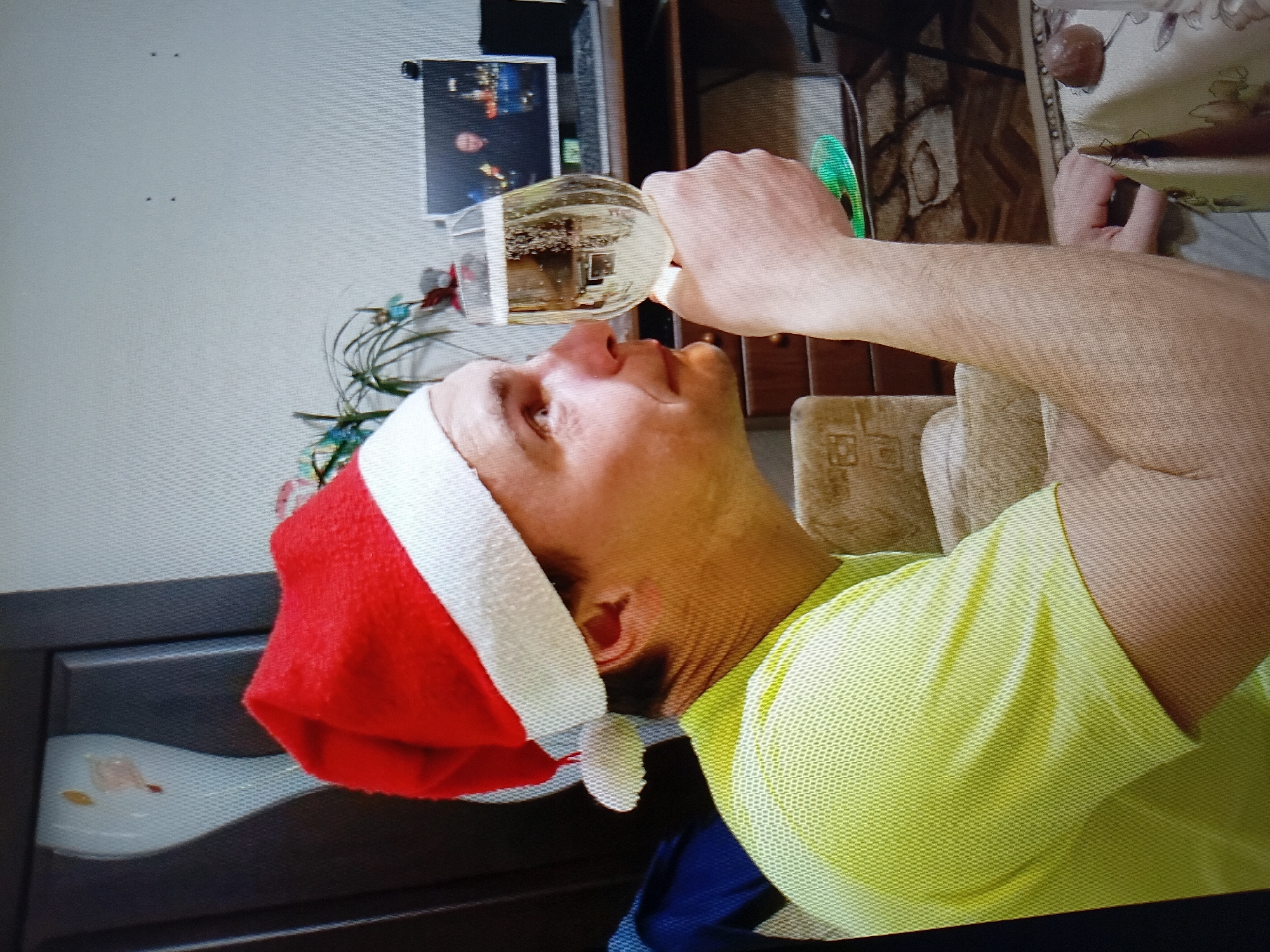 A man wearing a red Santa hat and a bright yellow t-shirt is holding a champagne glass to his lips. He is looking to his right, with his eyes open. In the background, a computer monitor displays two men, and a small green plant sits on a shelf. A dark-colored desk with drawers is visible behind the man. A light-colored couch is to his left. A portion of a patterned carpet is on the floor. A dark object resembling a chocolate is on a patterned tablecloth in the foreground. The setting appears to be an indoor residential environment, likely a living room or dining area. No specific landmarks or indicators for Tiraspol, Moldova are present in the image.