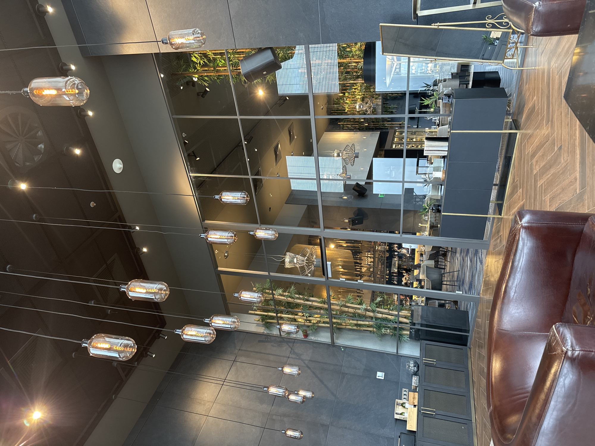 This image depicts a modern and elegantly designed interior space, likely a hotel lobby, lounge, or upscale restaurant, located in Sofia, Bulgaria. Numerous clear glass pendant lights with exposed filaments hang at varying heights from a dark grey ceiling, casting a warm, inviting glow.
The space is partitioned by large glass walls with a grid-like frame, creating reflections of the interior. To the right, a dark brown leather armchair or sofa, showing some wear, occupies the foreground. Beside it, a sleek black console table with gold-toned handles and accents is placed against a glass wall, behind which stands a tall, ornate gold-framed mirror.
Through the glass partitions, a decorative element resembling horizontal bamboo stalks is visible along a wall. Further beyond, other dining or lounge furniture and an intricate light fixture can be glimpsed. A large circular air vent or grille is noticeable on the dark ceiling in the upper-left. The flooring transitions from dark grey large-format tiles to a warm, herringbone-patterned wooden floor.
The overall ambiance is one of sophisticated tranquility, with no people actively present. The warm illumination suggests either evening hours or a deliberately intimate setting. Faintly visible through one of the glass panels, the text "TAITTINGER" can be discerned on a black surface, likely referencing a champagne brand. The clean lines, reflective surfaces, and strategic lighting contribute to a contemporary and luxurious feel in this Bulgarian establishment.