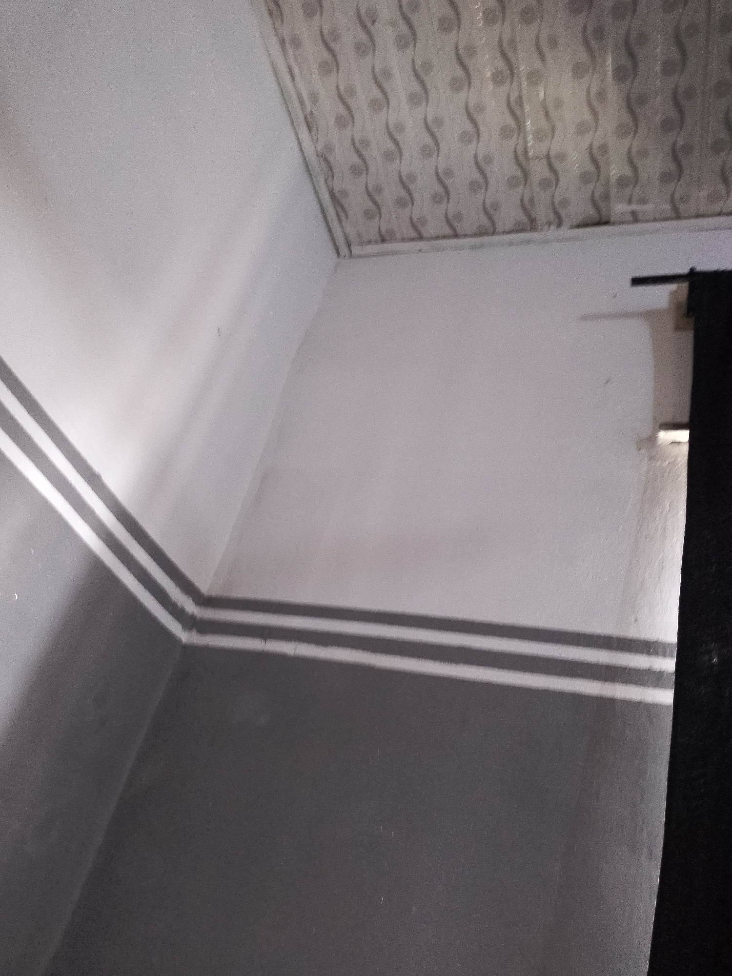 The image shows an interior view of a room with gray and white walls. The lower portion of the walls is painted dark gray, and the upper portion is white. There is a decorative stripe composed of two thin white lines running horizontally along the wall where the gray and white sections meet. The ceiling is covered with a textured material featuring a repeating wavy pattern in shades of gray and white. In the upper right corner, a dark object, possibly a curtain or a doorway, is visible, partially obscuring a light-colored wall. The lighting appears to be dim, casting shadows on the walls. The location is specified as Unknown, Unknown.