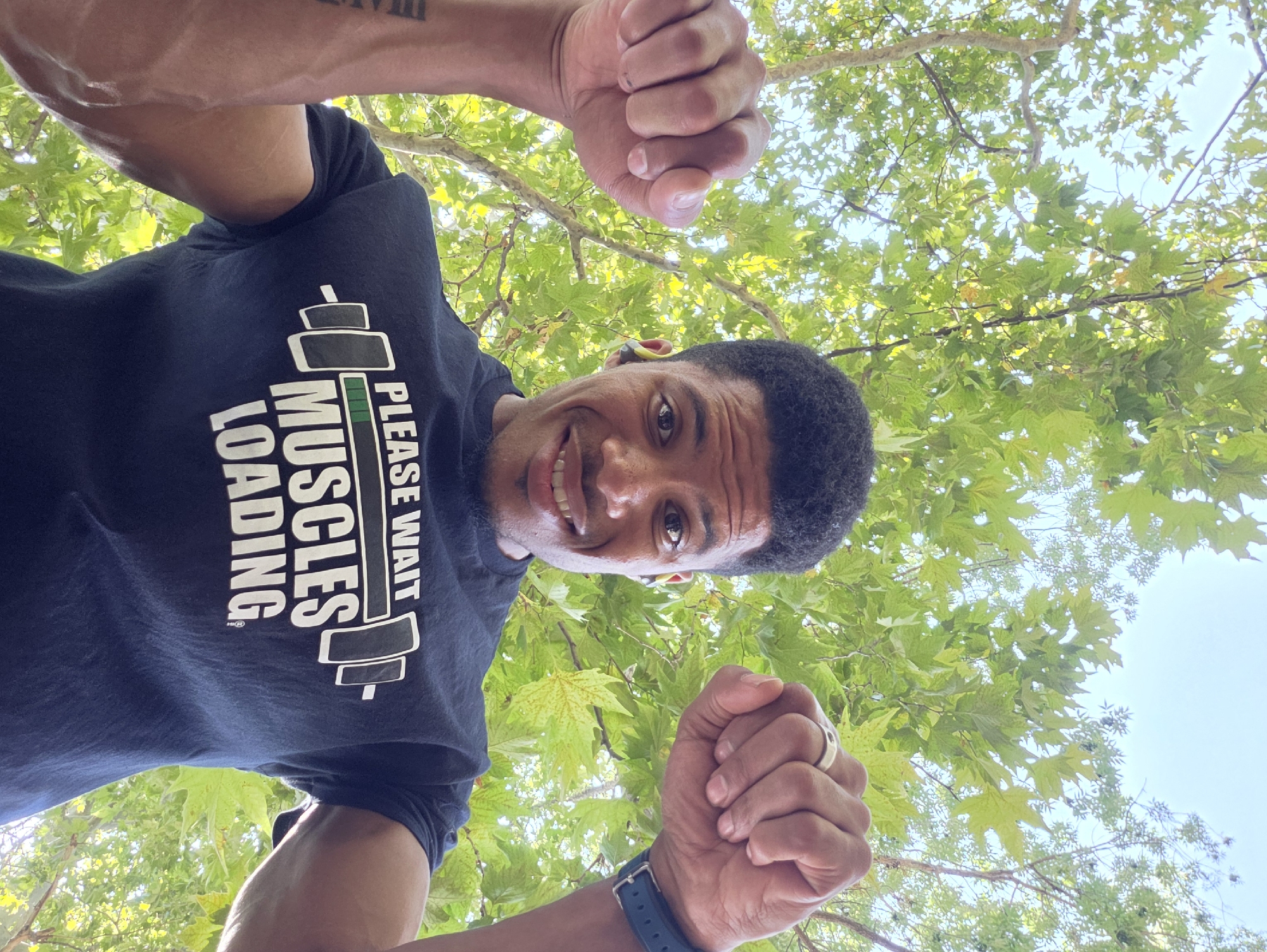 A man, viewed from a low angle, is looking up and smiling. He is wearing a dark blue t-shirt with the text "PLEASE WAIT MUSCLES LOADING" and an image of a barbell. He has short, dark, curly hair and is wearing wireless earbuds. His right arm is raised, and his fist is clenched. A ring is visible on his right ring finger, and a dark blue watch is on his left wrist. His left arm is also raised, with his fist clenched. The background consists of green leaves and a bright sky, suggesting an outdoor environment. The image does not provide any specific clues to indicate it was taken in the city of Varna, Bulgaria.