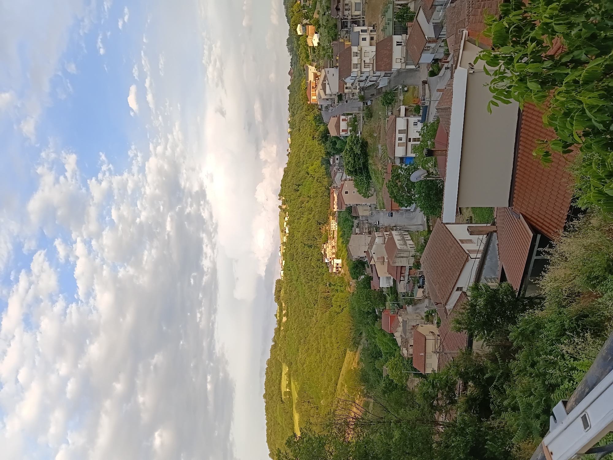 This is a high-angle, panoramic, eye-level shot of a small town nestled in a valley, with rolling green hills and a cloudy sky. The town appears to be Vallesaccarda, Italy, judging by the distinctive architecture and landscape. The houses are primarily constructed with light-colored stone and feature red tile roofs, typical of the region. The houses are clustered together, lining narrow, winding streets that snake through the town. Lush green trees and vegetation are abundant, surrounding the town and climbing up the slopes of the hills. The sky is partly cloudy, with patches of blue peeking through, suggesting it might be late afternoon or early evening. There are no people visible in the image, nor any discernible actions or interactions. The scene is serene and peaceful, capturing the essence of a quiet Italian village.