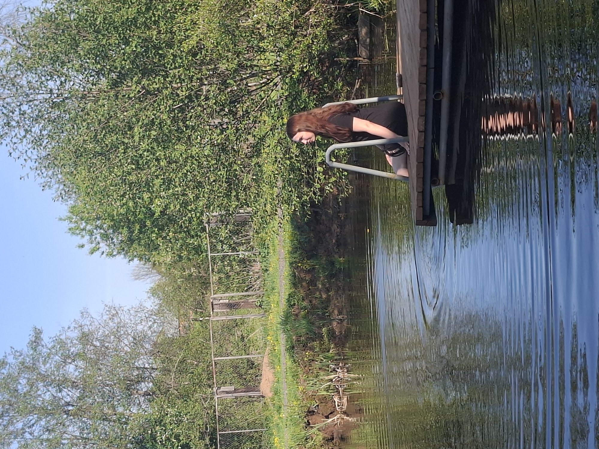 The scene depicts an unidentified woman standing on a dock extending into a body of water, likely a pond or lake. She is gripping the metal ladder of the dock and appears to be looking down. The woman has dark hair, a dark-colored short-sleeved shirt, and dark bottoms.
The dock is constructed of wood with a metal ladder providing access to the water. The water's surface is rippled with reflections of the surrounding trees and dock structure.
The backdrop features lush green trees. A chain-link fence runs along the bank. The sky is partly cloudy.
The location is potentially Malyi Bukor, Russia, but the image contains no indicators to confirm the city. The environment suggests a park or natural recreational area.