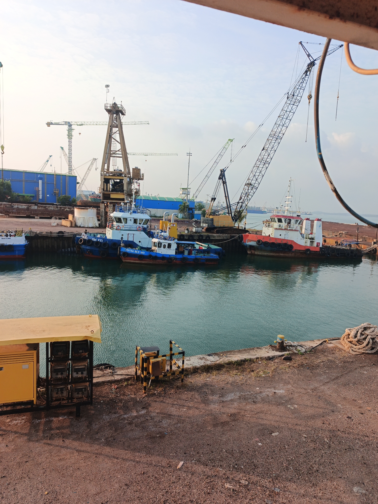 The image depicts a harbor scene in Batam, Indonesia, likely within a shipyard or industrial port area, characterized by heavy machinery and vessels. In the foreground, a rough, reddish-brown ground surface is visible, with a yellow industrial generator and electrical meters on the left. Further back, a concrete pier juts out into a body of calm, turquoise water.
Dominating the water are several boats. Three tugboats are moored side-by-side. The leftmost is mostly out of frame, but appears white with blue accents. The middle tugboat is blue with white superstructure and has "SM XXI" painted on its side. The rightmost tugboat is smaller and painted blue and white. Further to the right, a larger vessel with a red and white hull, likely a tug or workboat, is also docked.
The background is filled with large industrial cranes, including a prominent tall tower crane in the center and a massive lattice boom crane on the right. Several other cranes, some green and others more industrial in appearance, are scattered throughout the scene, indicating active construction or loading operations. Large blue industrial buildings are visible behind some of the cranes. The sky is overcast with a bright, diffused light, suggesting it is either early morning or late afternoon. The overall impression is one of industrial activity and maritime operations in a busy port.