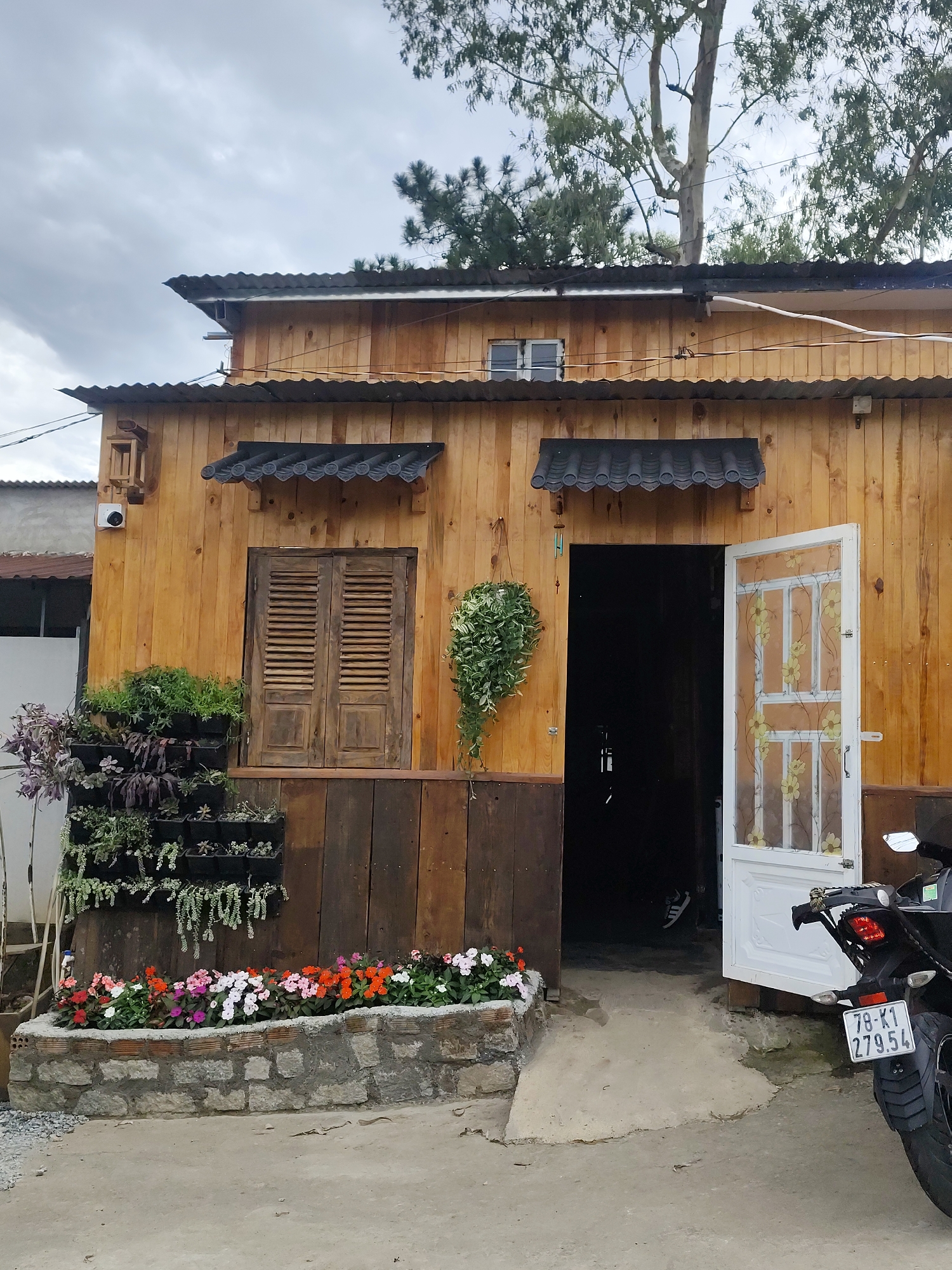 The image depicts a charming, rustic wooden house in Da Lat, Vietnam. The house has a corrugated metal roof and is adorned with various plants and flowers. A vertical garden is mounted on the wooden wall, filled with lush green succulents and trailing plants. In front of the house, a low stone wall encloses a vibrant flower bed bursting with colorful blooms in shades of pink, white, orange, and red.
To the right of the entrance, a white door with decorative floral patterns is open, revealing a dark interior. A black motorcycle with a license plate reading "78-K1 279.54" is parked beside the doorway, suggesting it's a mode of transportation for someone associated with the house. A single pair of white sneakers is visible inside the dark doorway, hinting at a person's presence.
Above the wooden shutters of a window, a small, decorative awning with curved tiles adds a traditional touch. Hanging from the side of the house is a small, wind chime. The overall scene evokes a peaceful and idyllic atmosphere. The lighting suggests it is daytime, possibly overcast due to the muted light and cloudy sky visible at the top of the frame. The presence of trees and the architectural style point towards a location in Vietnam, consistent with the Da Lat context.