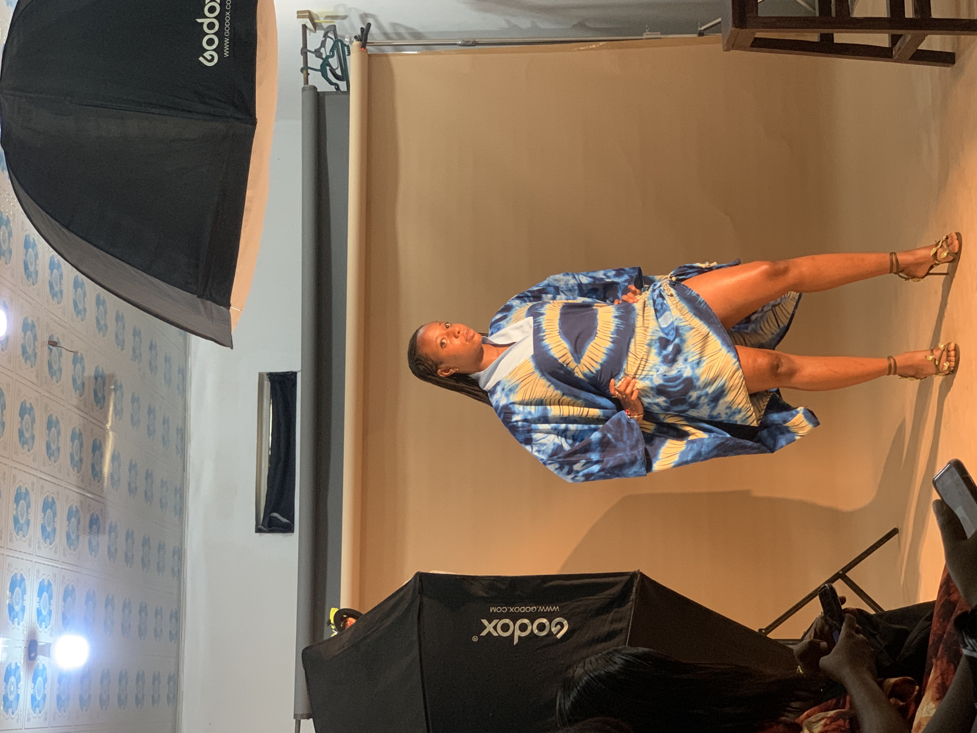 The image appears to be a studio shot of a woman posing in front of a neutral background. She is wearing a blue and white patterned dress and sandals with gold embellishments. She is posing with her legs extended. In the foreground, there are photography lights and some onlookers with mobile phones. The location appears to be an indoor studio, potentially in Gura Top, Nigeria, although there is no definitive information to confirm the city. The environment is illuminated by studio lighting.