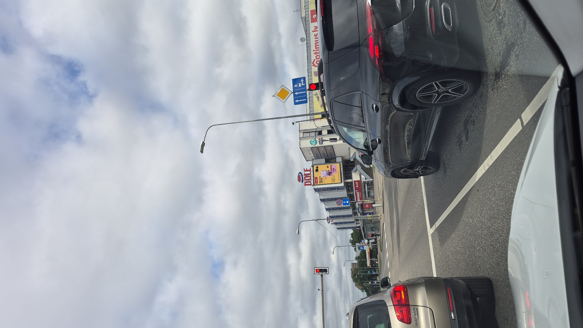 The image shows a view from a car looking out at a city street in Riga, Latvia. The sky is overcast with clouds. In the foreground, the hood of the car from which the photo is taken is visible. To the left, the rear of a tan SUV is stopped at a traffic light. To the right, a black SUV is also stopped.
The street ahead shows multiple lanes of traffic and a crosswalk. On the right side of the street, there is a large building with signage that reads "DOLE" and "Optimus lv". There are several traffic lights visible, with one showing red in the middle distance. There are also streetlights and various road signs. A bus stop shelter is visible on the left side of the street, with a few people near it. The overall scene depicts a typical urban environment at a traffic intersection.