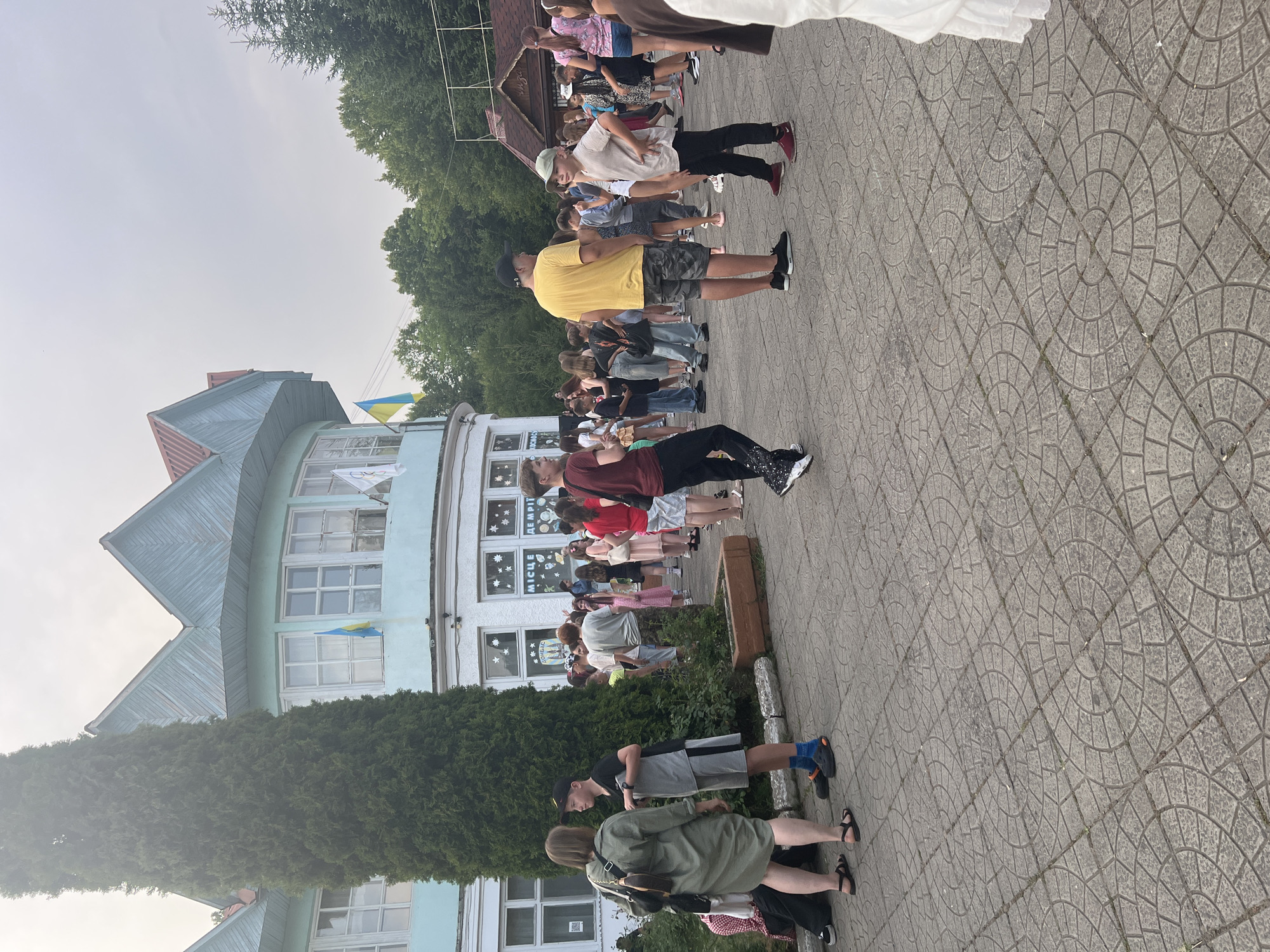 The image depicts an outdoor scene in front of a building with a light blue facade and a distinctive red-trimmed, multi-peaked roof. Ukrainian flags are visible on the building. A tall, dark green evergreen tree stands prominently to the left of the building. The ground is paved with interlocking circular stone patterns. A crowd of people, including adults and children, are gathered in the paved area.
In the foreground, a woman in a green dress and a boy in a grey t-shirt and grey shorts are interacting. The woman has a dark purse slung over her shoulder. The boy is wearing a black baseball cap and blue socks. To their right, a group of young people are standing. A boy in a yellow t-shirt and camouflage shorts is facing right, and a boy in a light beige t-shirt and black pants is facing left. Further back, more people are milling about, some standing in groups, others walking. The overall impression is of a public gathering or event. The presence of the Ukrainian flags suggests this location is in Ukraine. Based on the request to include the city of Сонячне, Ukraine, this scene is understood to be situated there.