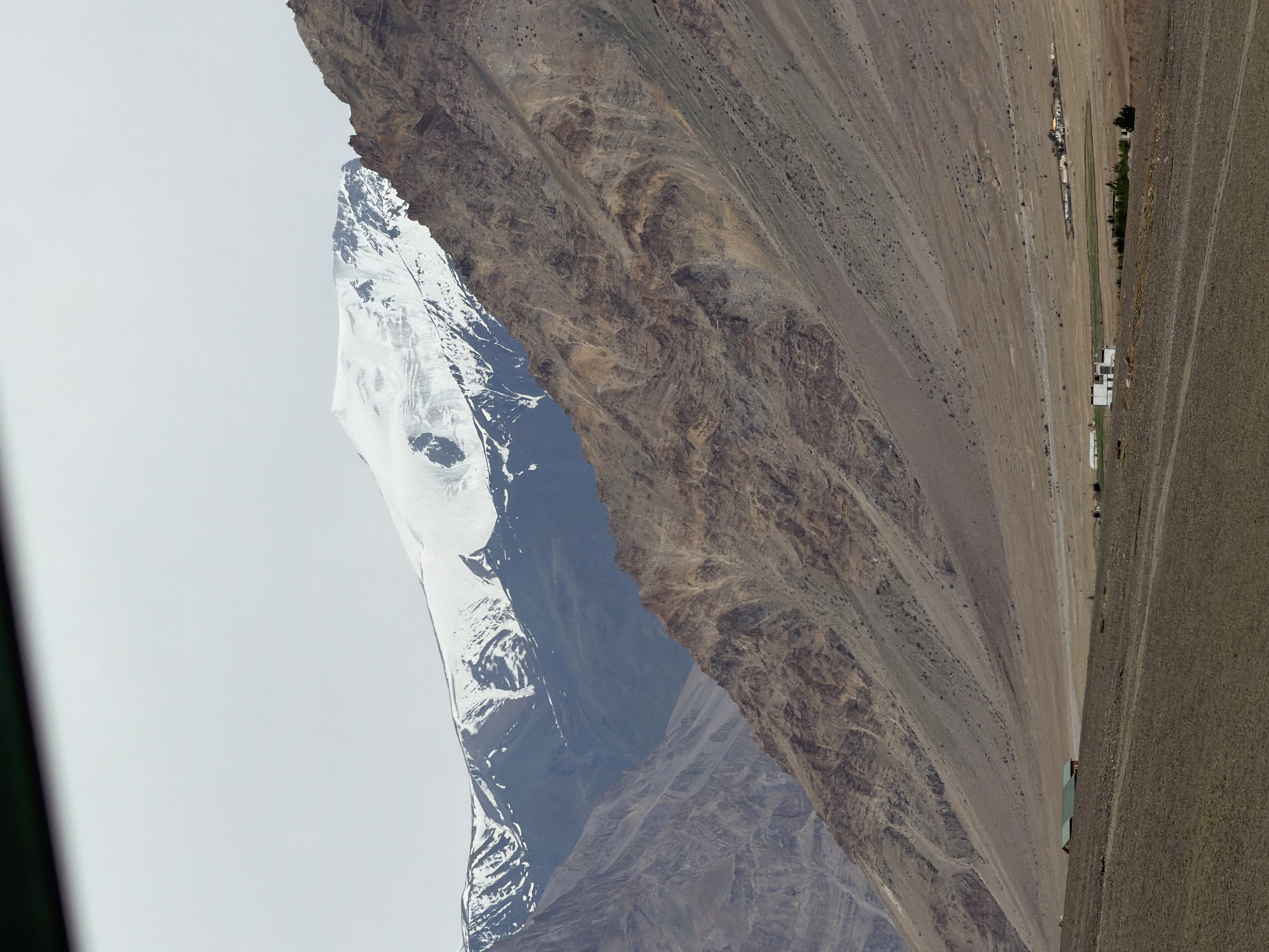 The image shows a vast, arid mountainous landscape under a cloudy sky. Dominating the background are towering, snow-capped mountains, their peaks shrouded in white. The slopes of the mountains are a mix of barren, rocky terrain with visible geological strata and patches of scree. In the mid-ground, a vast, flat valley floor stretches out, sparsely dotted with buildings. Two small, rectangular structures with green roofs are visible on the left side of the valley floor. Further to the right, a cluster of white buildings forms a small settlement, and beyond that, a few more scattered structures are discernible. The foreground consists of a dry, dirt track or road, with a rough, uneven surface. The overall impression is one of a remote, rugged, and sparsely populated mountainous region, characteristic of a high-altitude area like Zangla, India.
